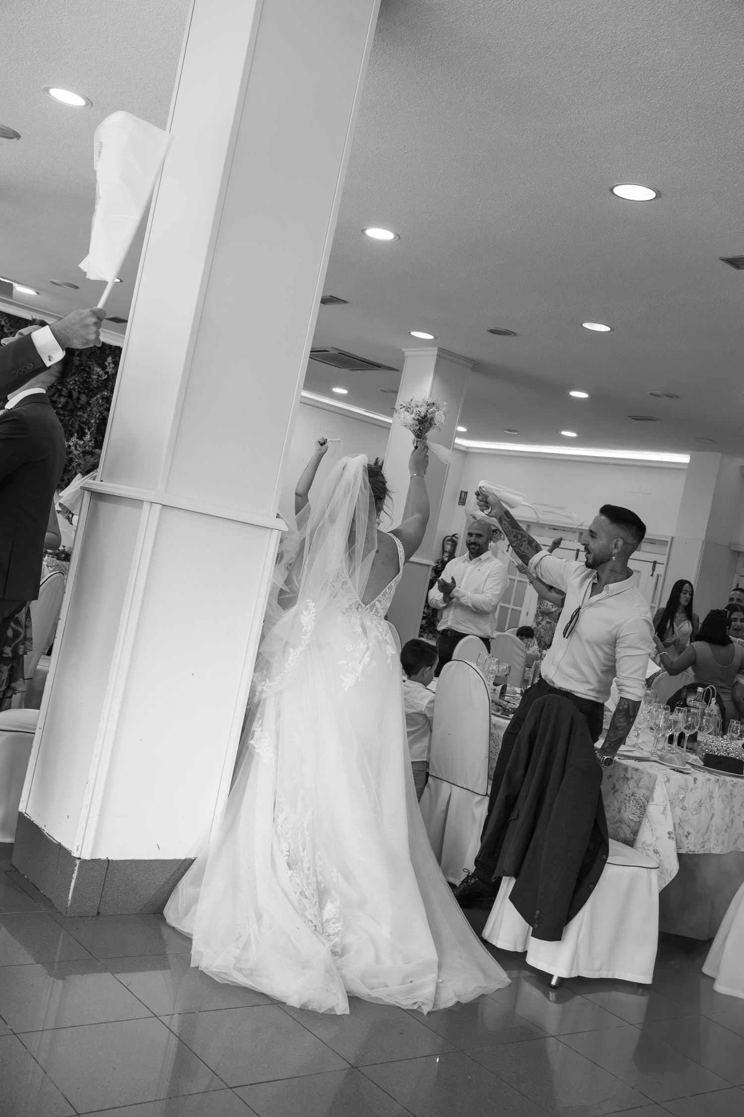 Black and white photo of a bride in a lace wedding gown raising her bouquet while guests dance and celebrate at a banquet reception.