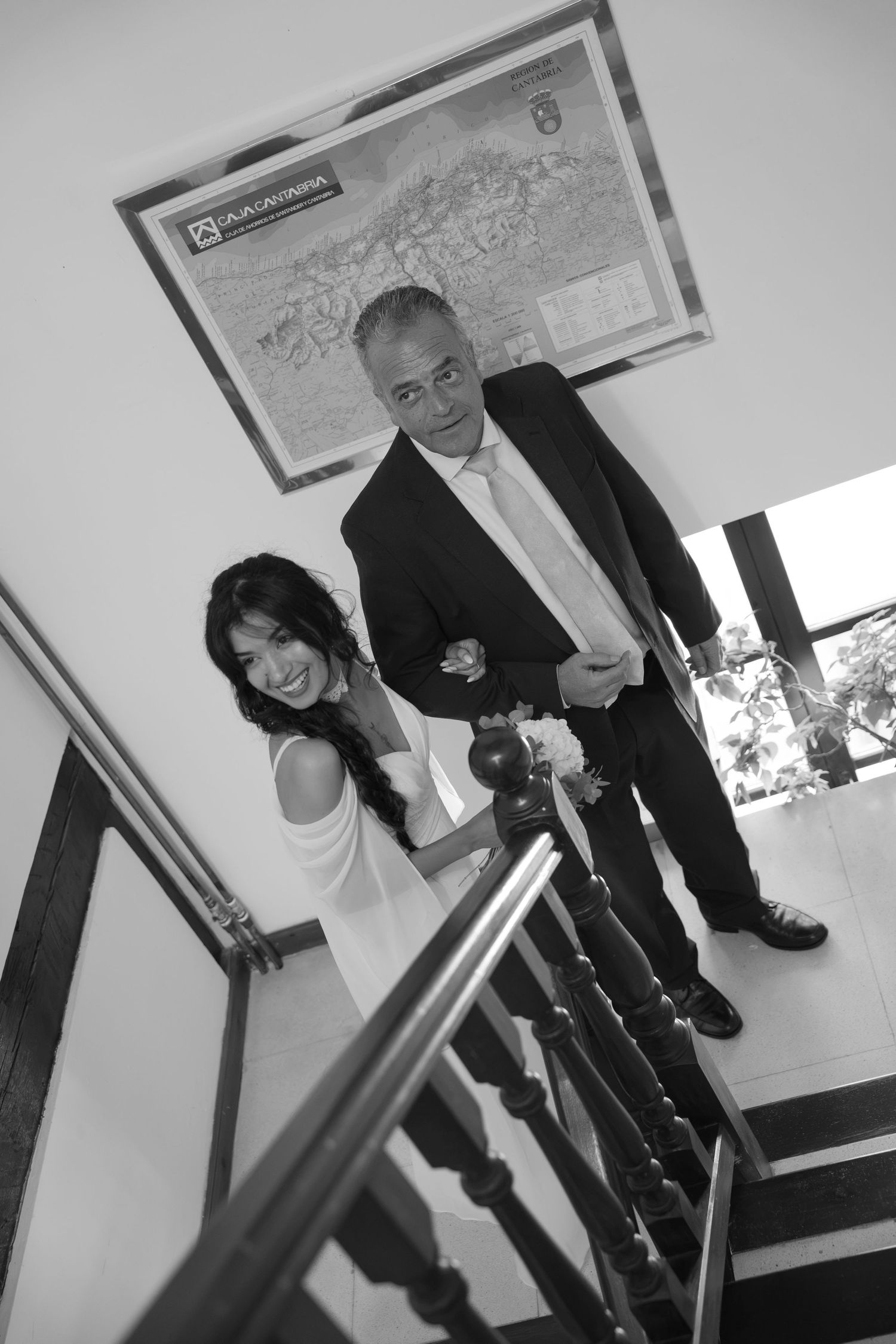 Black and white photo of a smiling bride holding a bouquet beside a man in a suit on a staircase with a framed map on the wall.