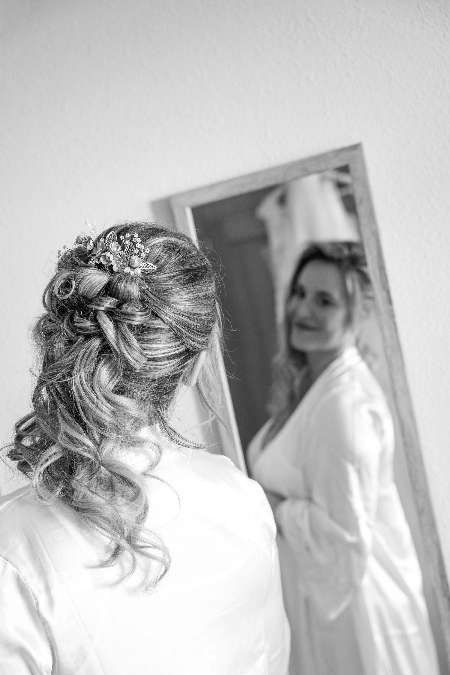 Black and white photo of a bride in a satin robe with a floral hair accessory and curled updo, smiling at her reflection in a mirror.
