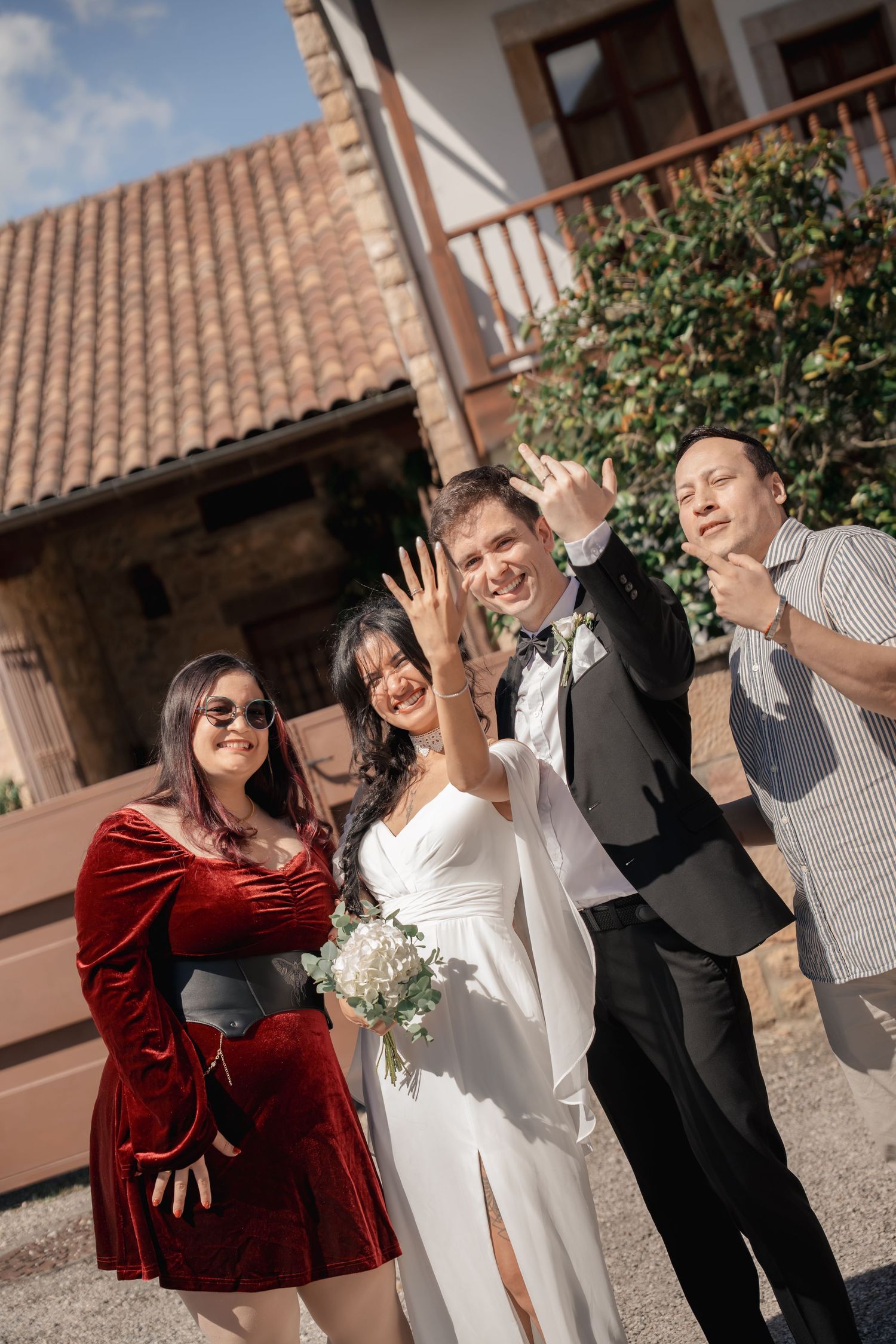Bride in a white dress holding a bouquet poses with smiling wedding guests outside a rustic stone building with a terracotta roof.