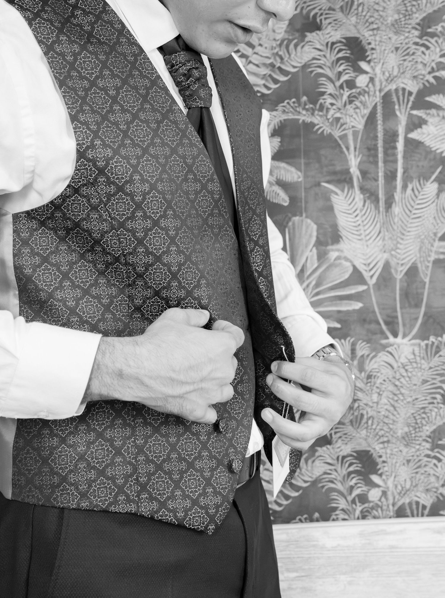 Groom in a patterned vest, white shirt and ruched cravat adjusts his waistcoat in front of fern leaf wallpaper in black and white.