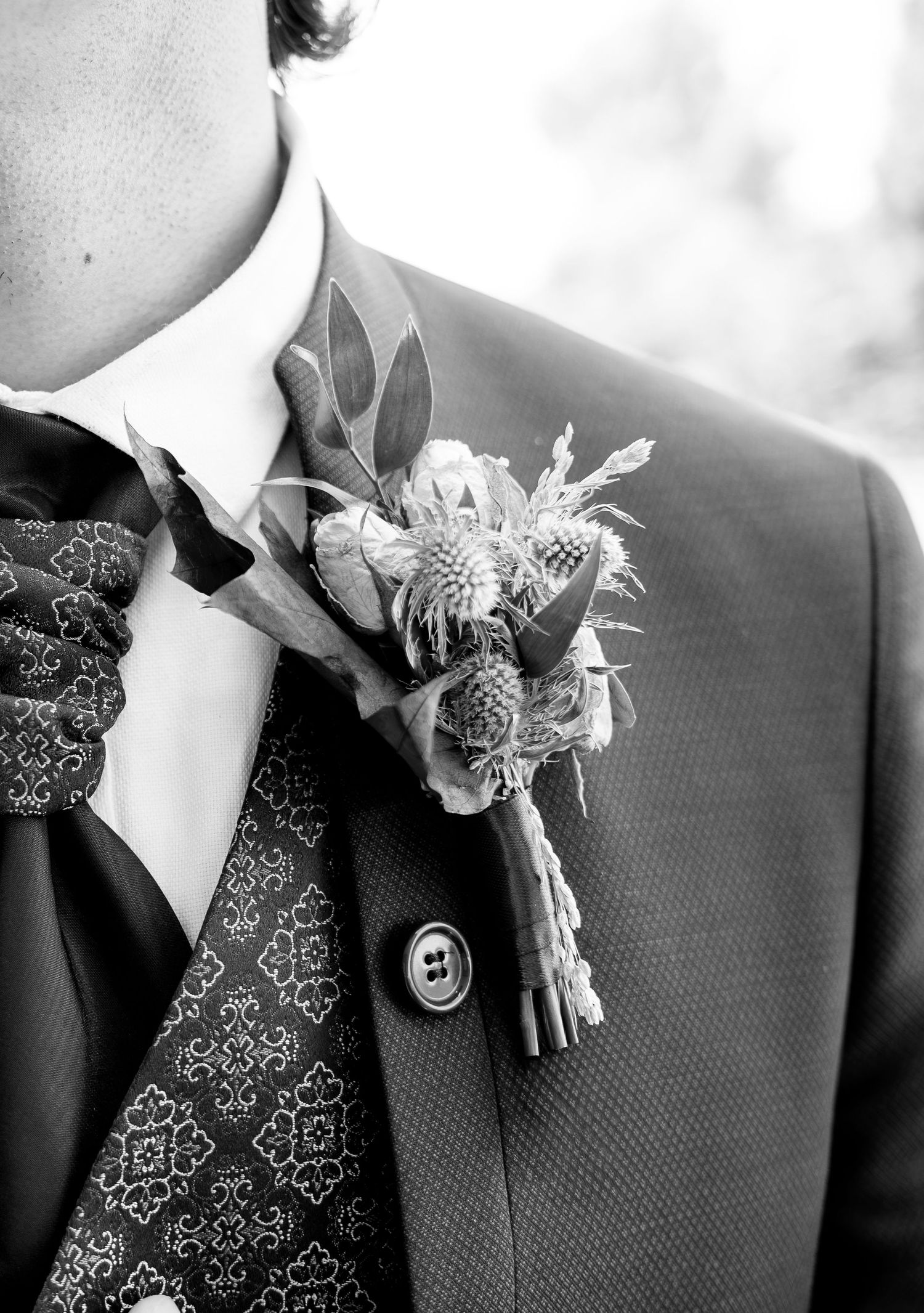 Black and white close-up of a groom's suit with a thistle and foliage boutonniere, patterned waistcoat and cravat.