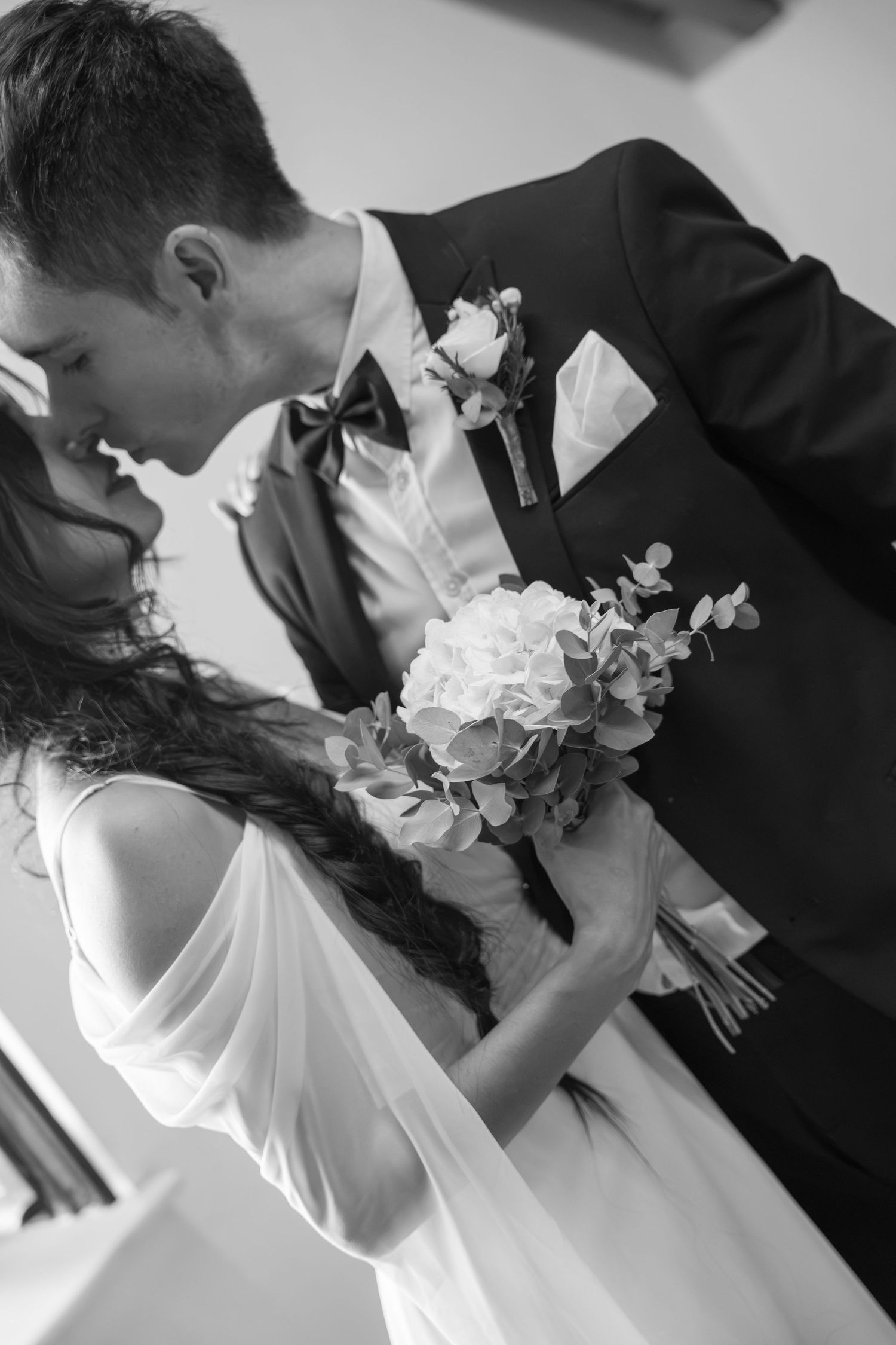 Black and white photo of a bride holding a bouquet and groom in a suit sharing an intimate moment on their wedding day.