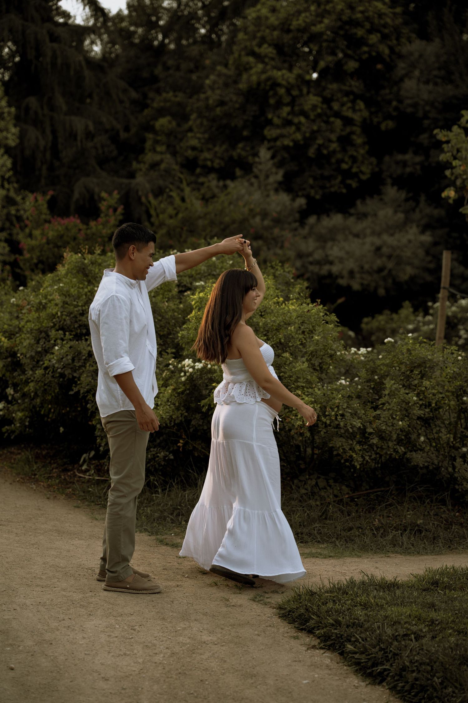 Couple sharing romantic dance on garden path, woman in white dress being twirled by partner at sunset.