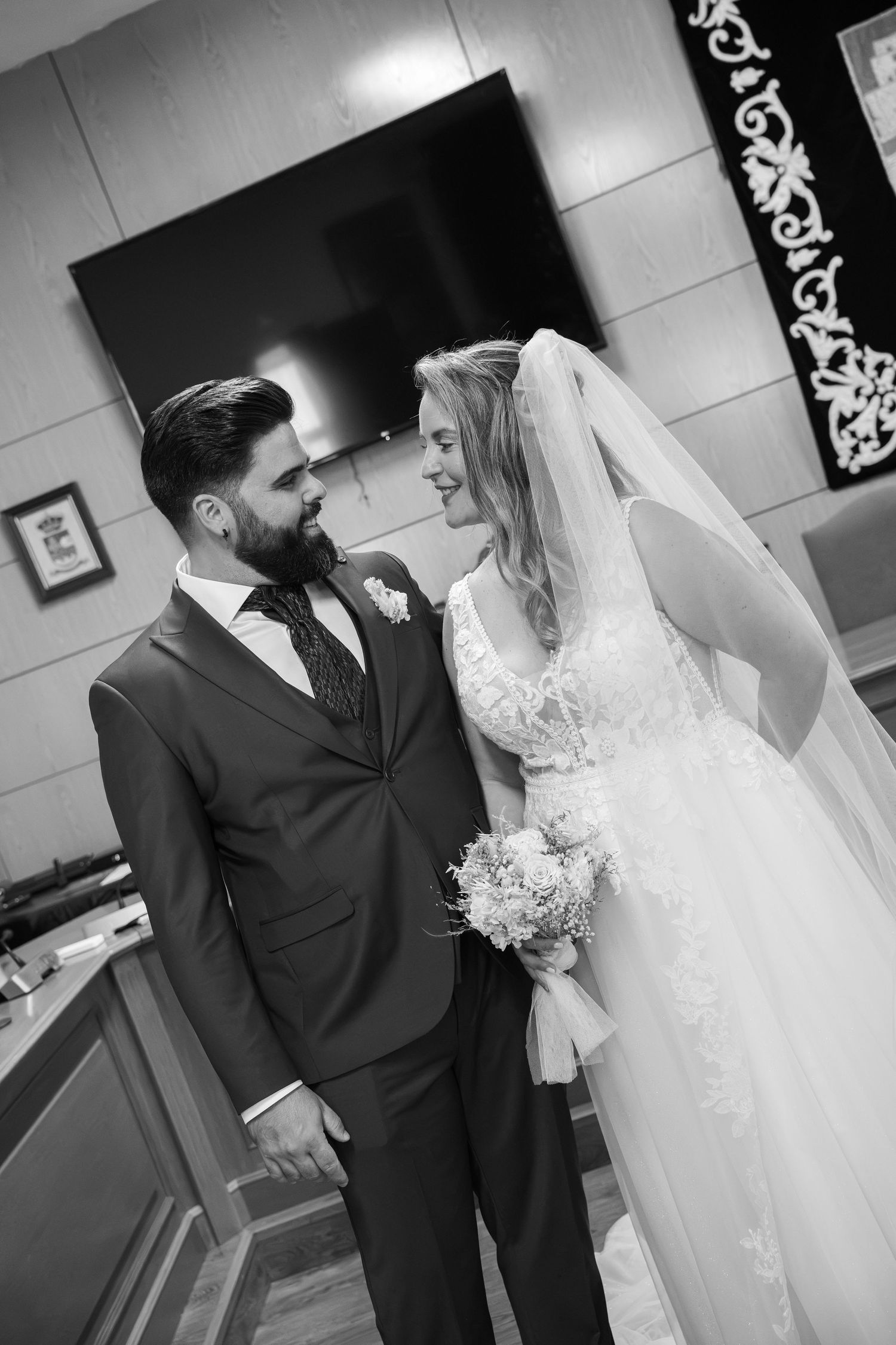 Bride in a lace gown and veil smiles at a bearded groom in a suit and tie during a wedding ceremony, in black and white.
