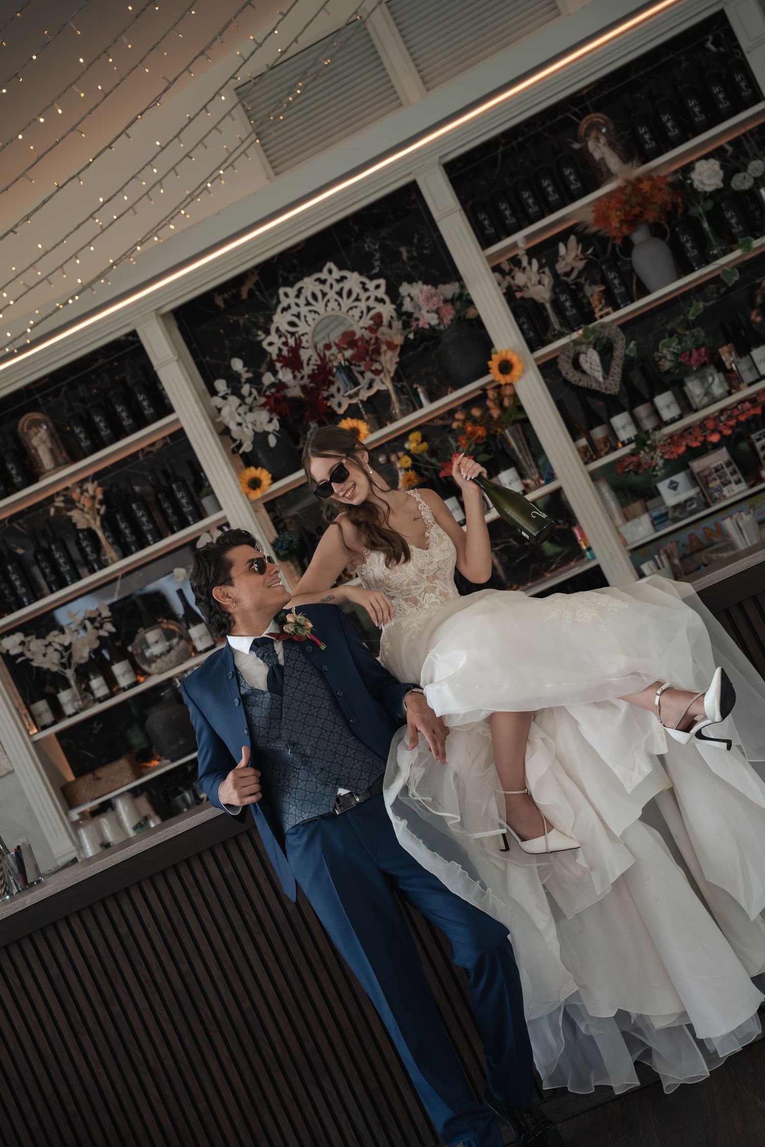 Groom in blue suit lifts bride in white gown at a decorated bar, both wearing sunglasses, celebrating their wedding day.