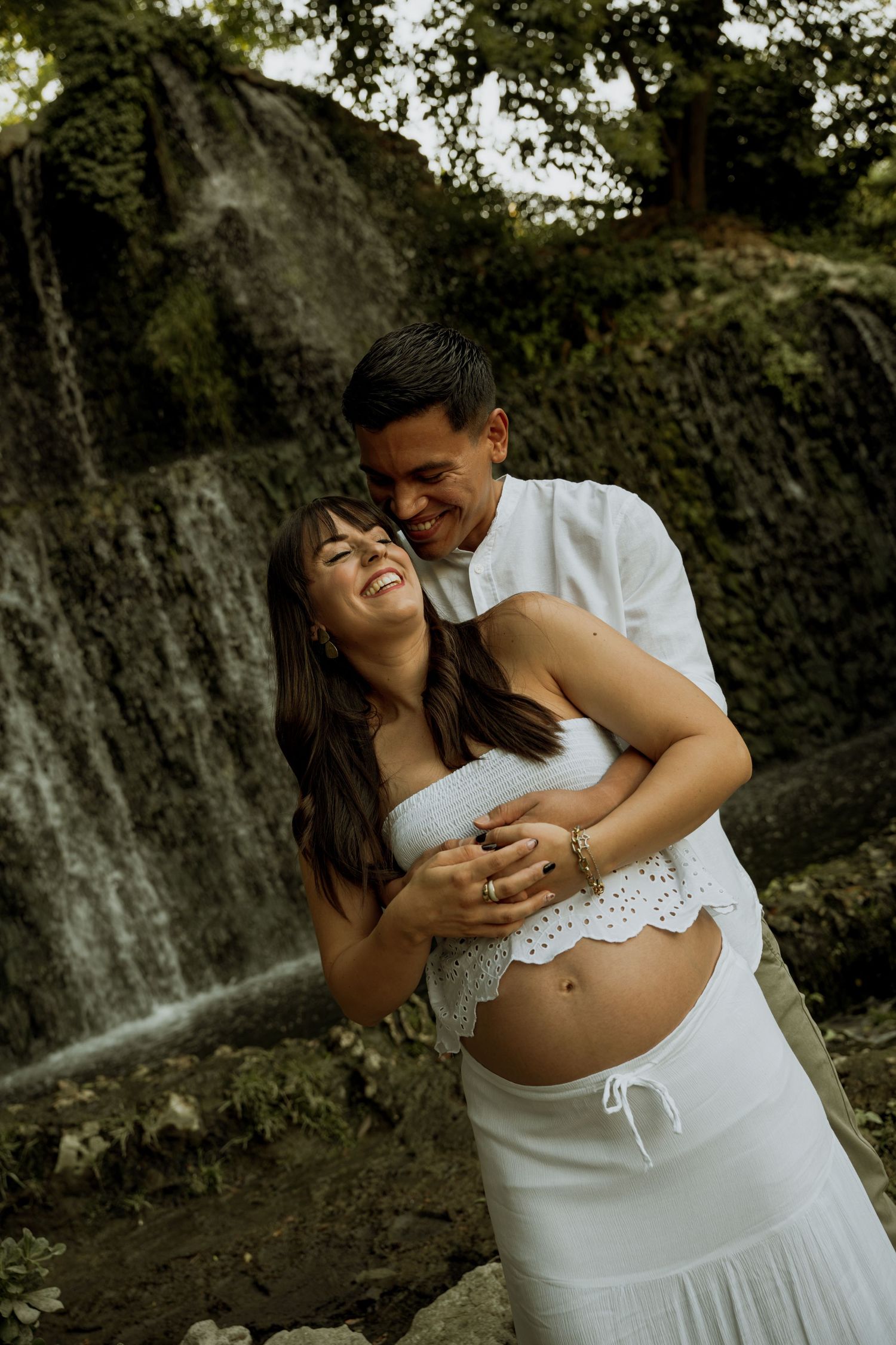 Happy expecting couple embracing in front of a waterfall, celebrating their maternity photoshoot.