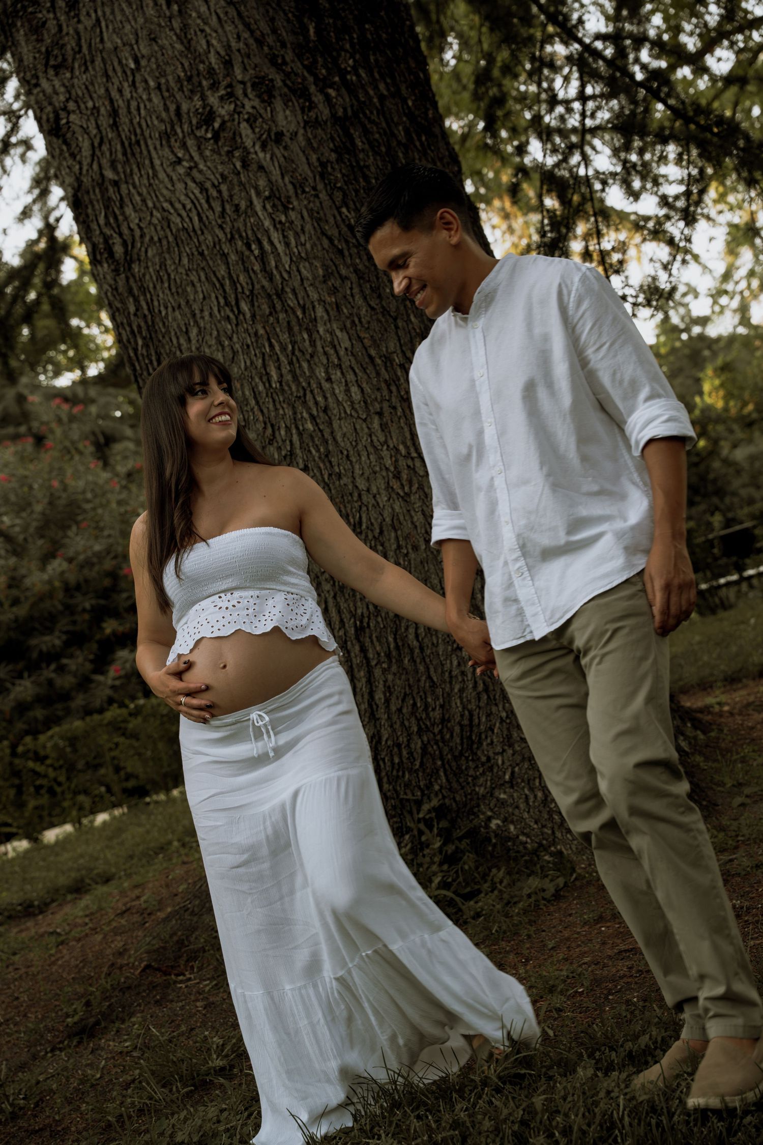 Pregnant woman in white outfit holding baby bump while smiling with partner leaning against large tree.