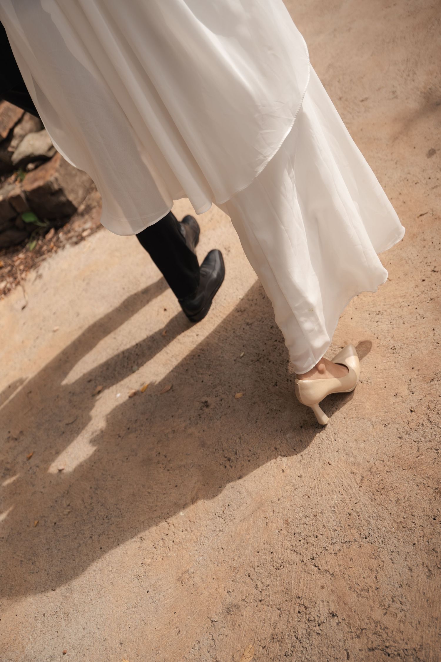 Bride in a flowing white dress and gold high heels walks beside a groom in black shoes and trousers on a sunlit dirt path.