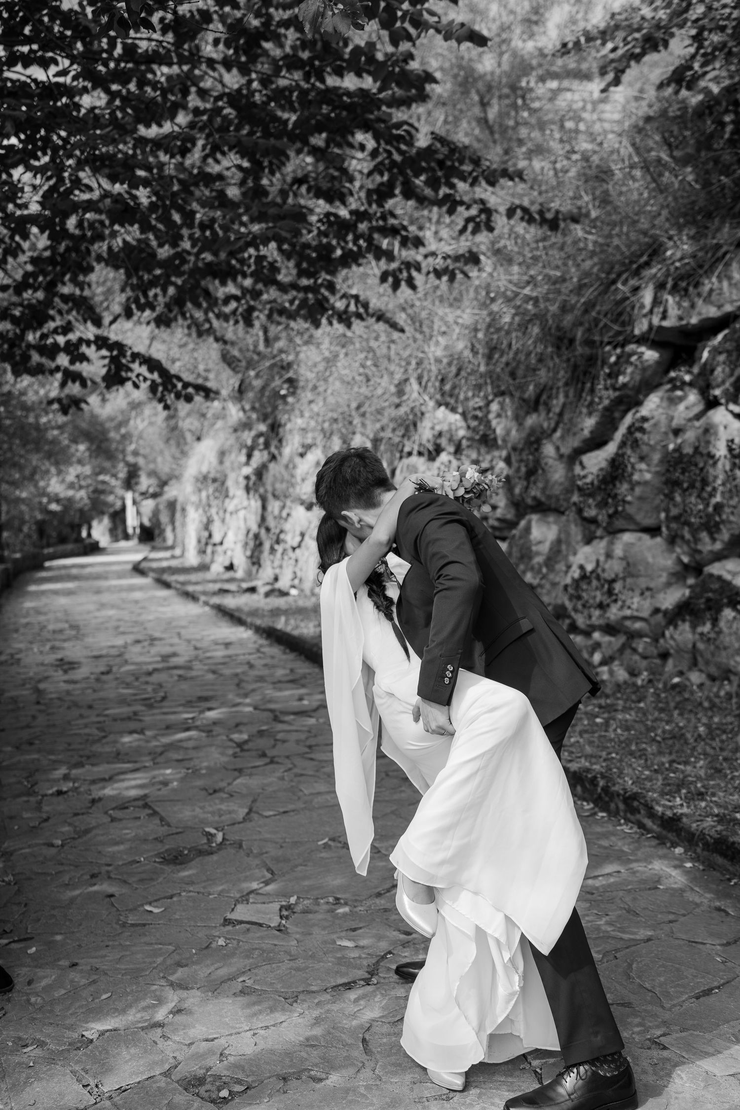 Black and white photo of a groom dipping and kissing his bride in a flowing white gown on a stone path lined with trees.