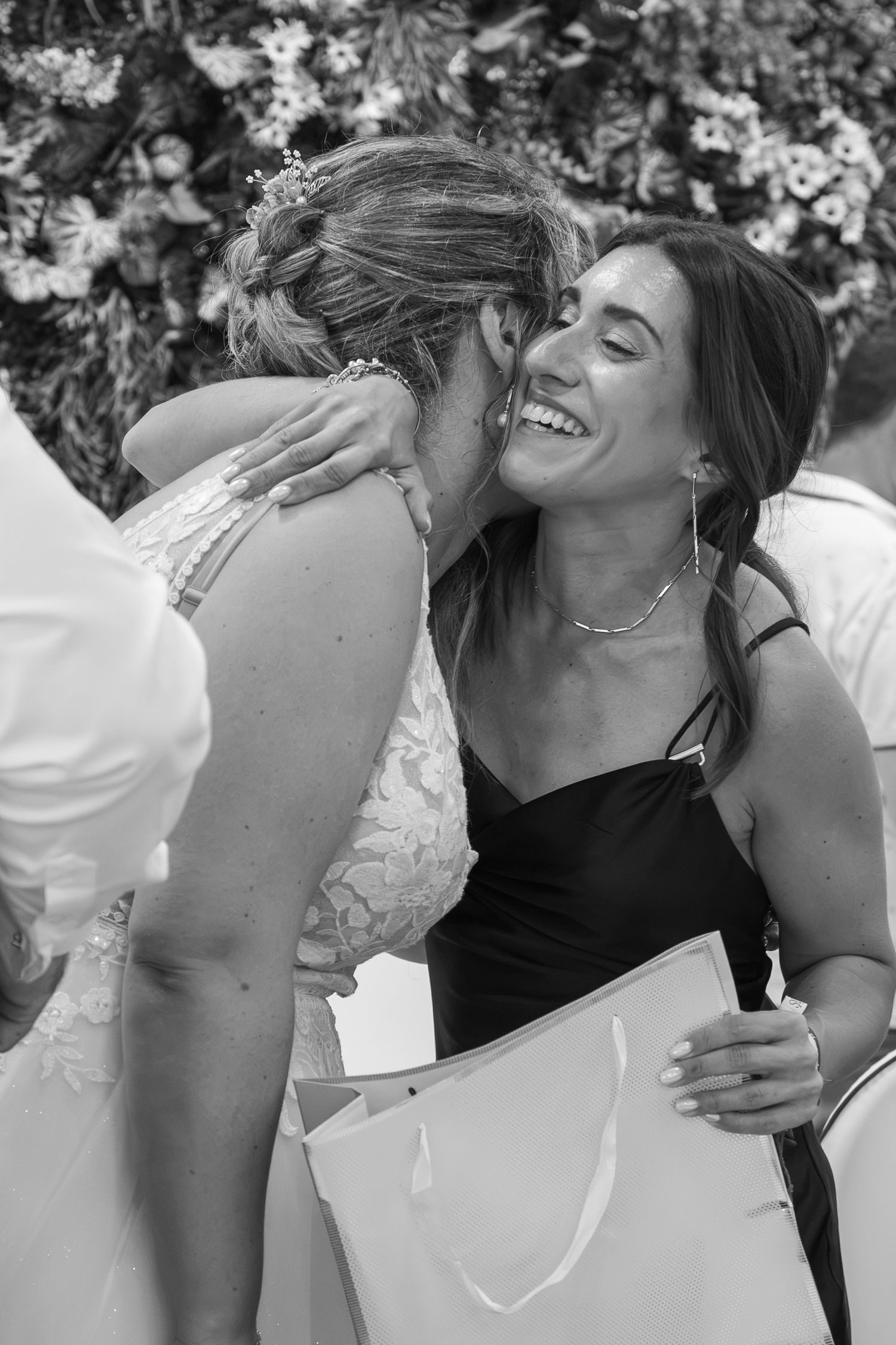 Black and white photo of two women laughing and hugging at a wedding, one in a lace gown, the other in a black dress holding a gift bag.