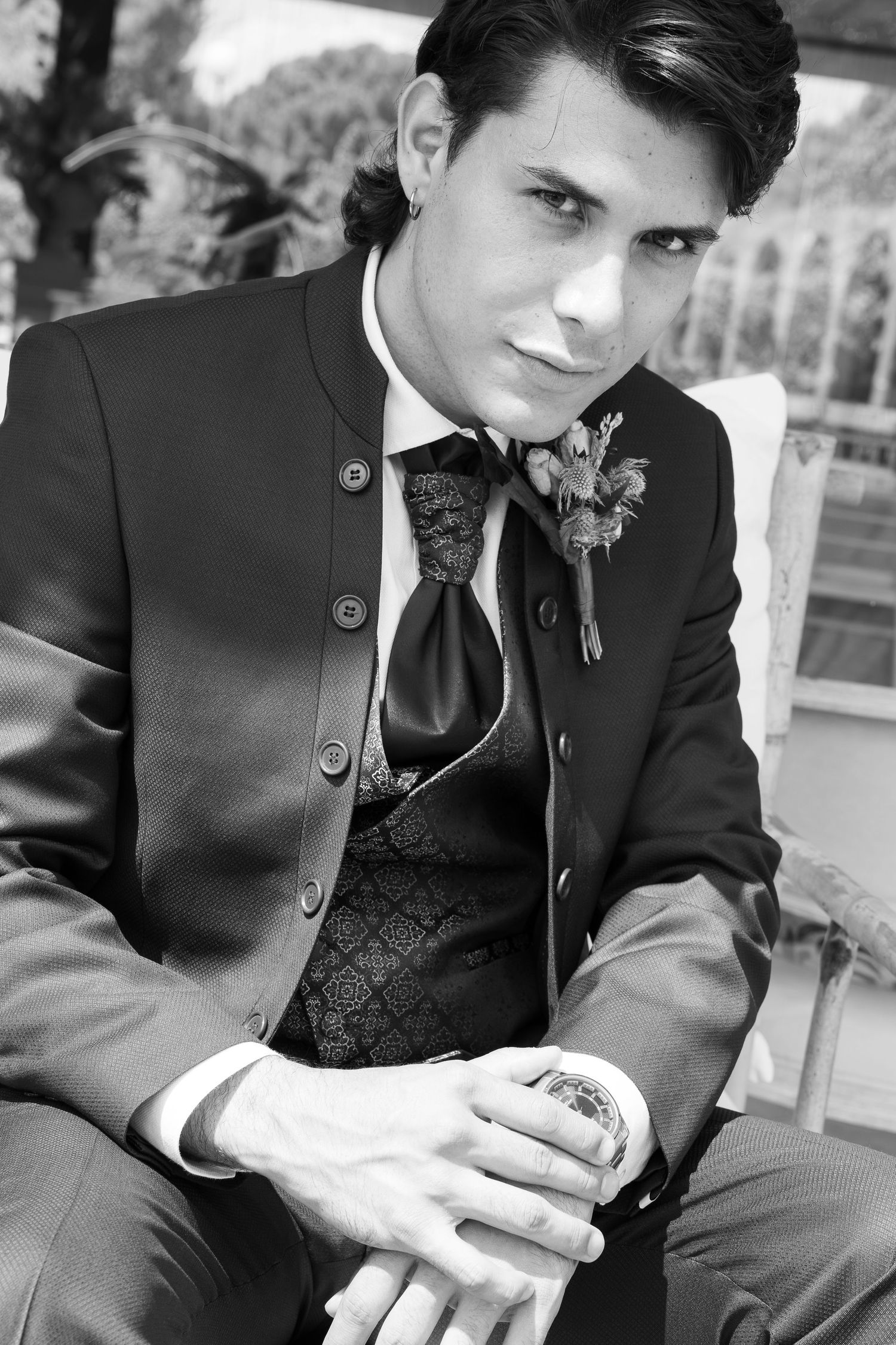 Black and white portrait of a groom in a buttoned jacket, patterned vest, bow tie and floral boutonniere, seated with hands clasped.