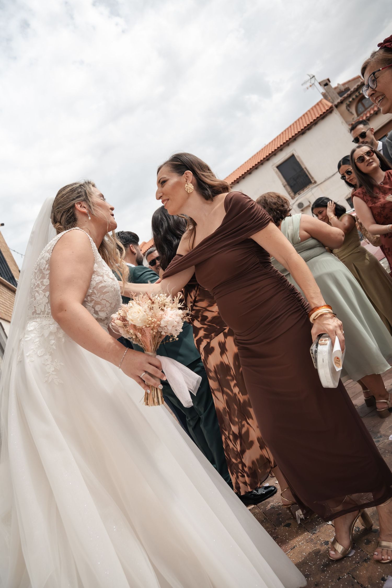A bride in a white lace gown holding a bouquet embraces a smiling guest in a brown dress outdoors while other wedding guests gather behind.