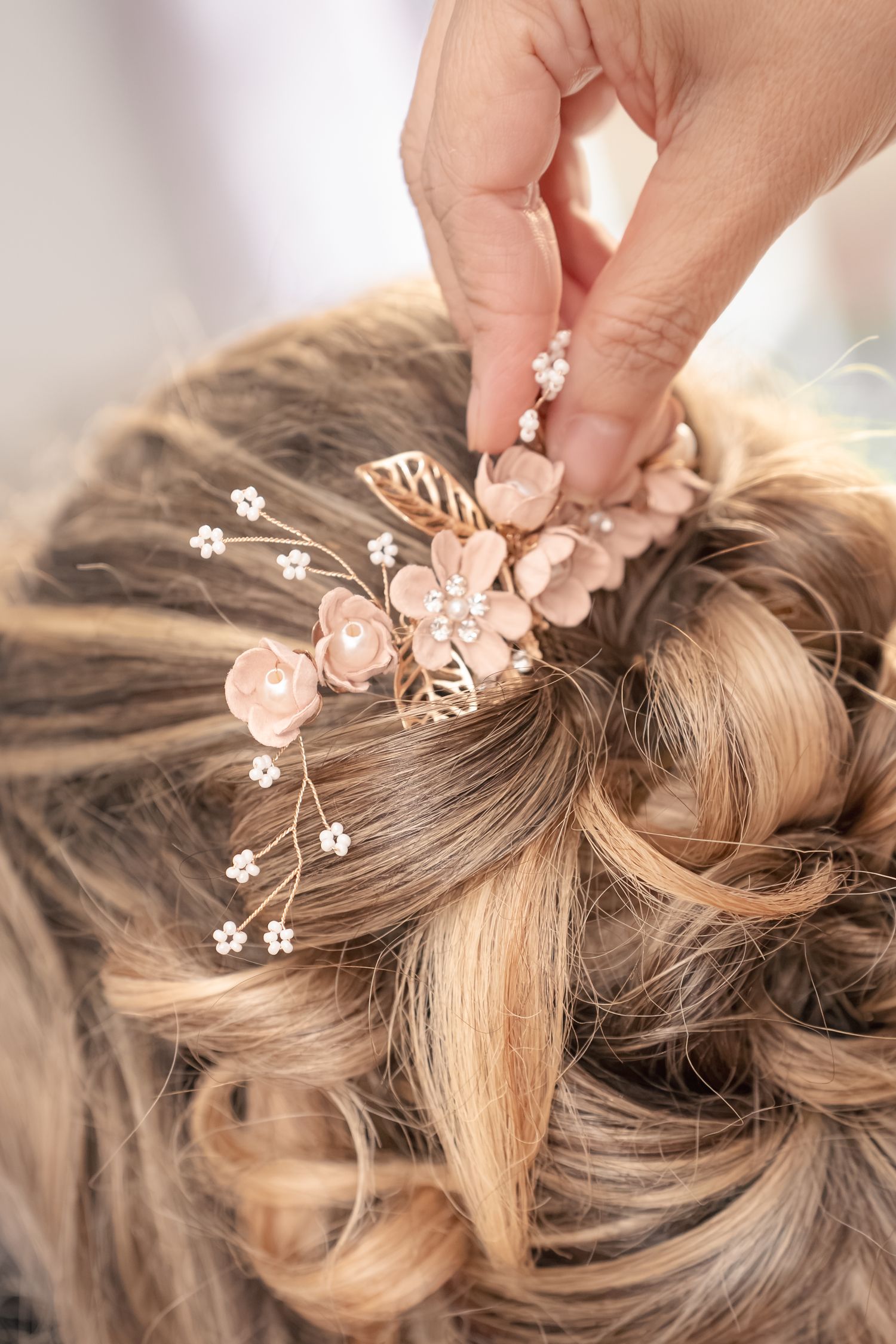 Hand placing a delicate blush pink floral hair pin with pearls and gold leaves into a curled blonde updo for a wedding hairstyle.