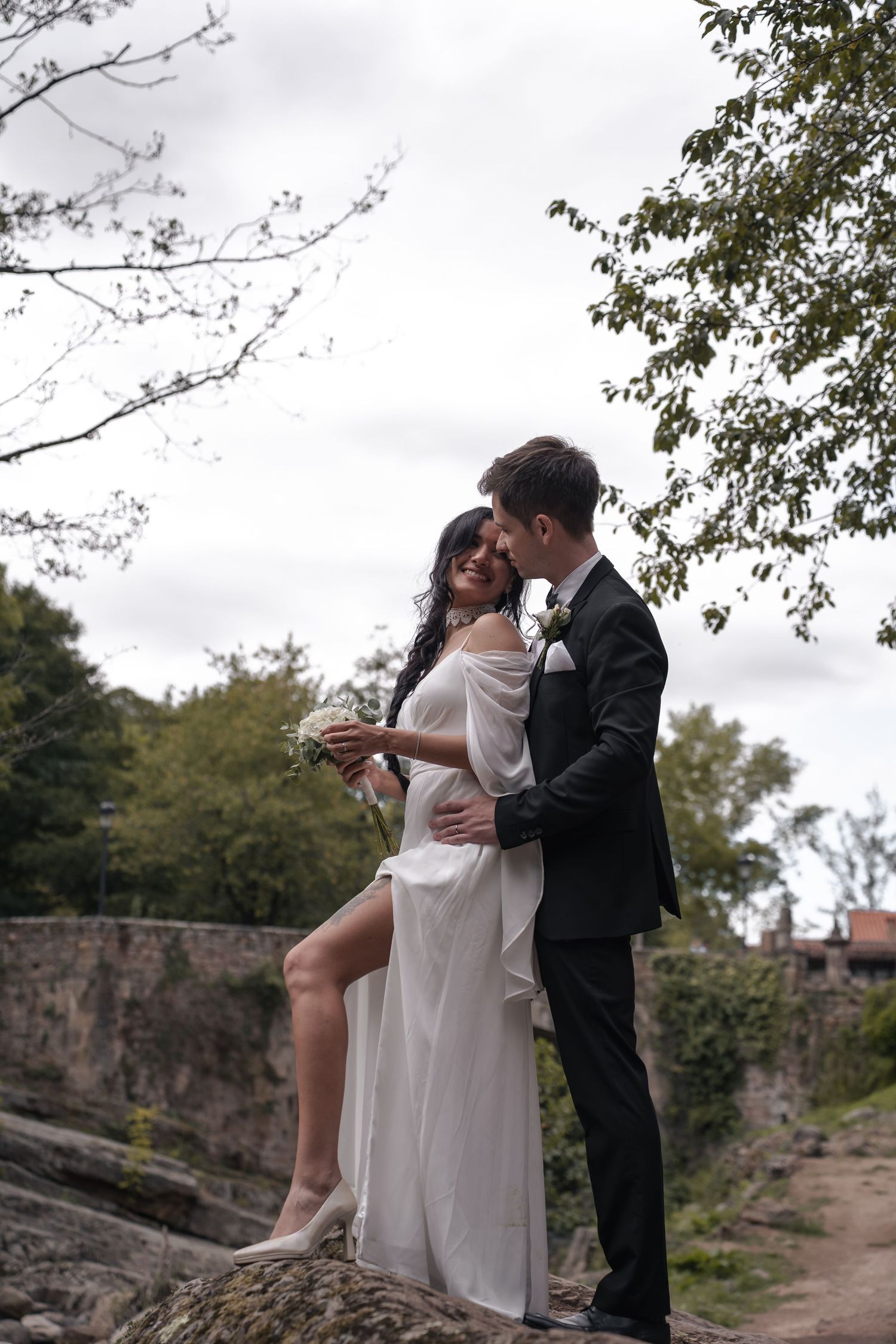 A bride and groom share a romantic embrace outdoors on rocks, surrounded by lush trees on their wedding day.