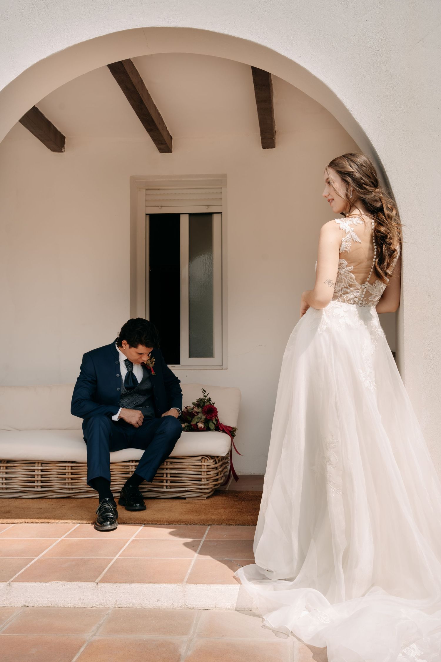 Bride in a lace-back wedding gown stands under a white arch while the groom in a navy suit sits on a wicker bench, head bowed.