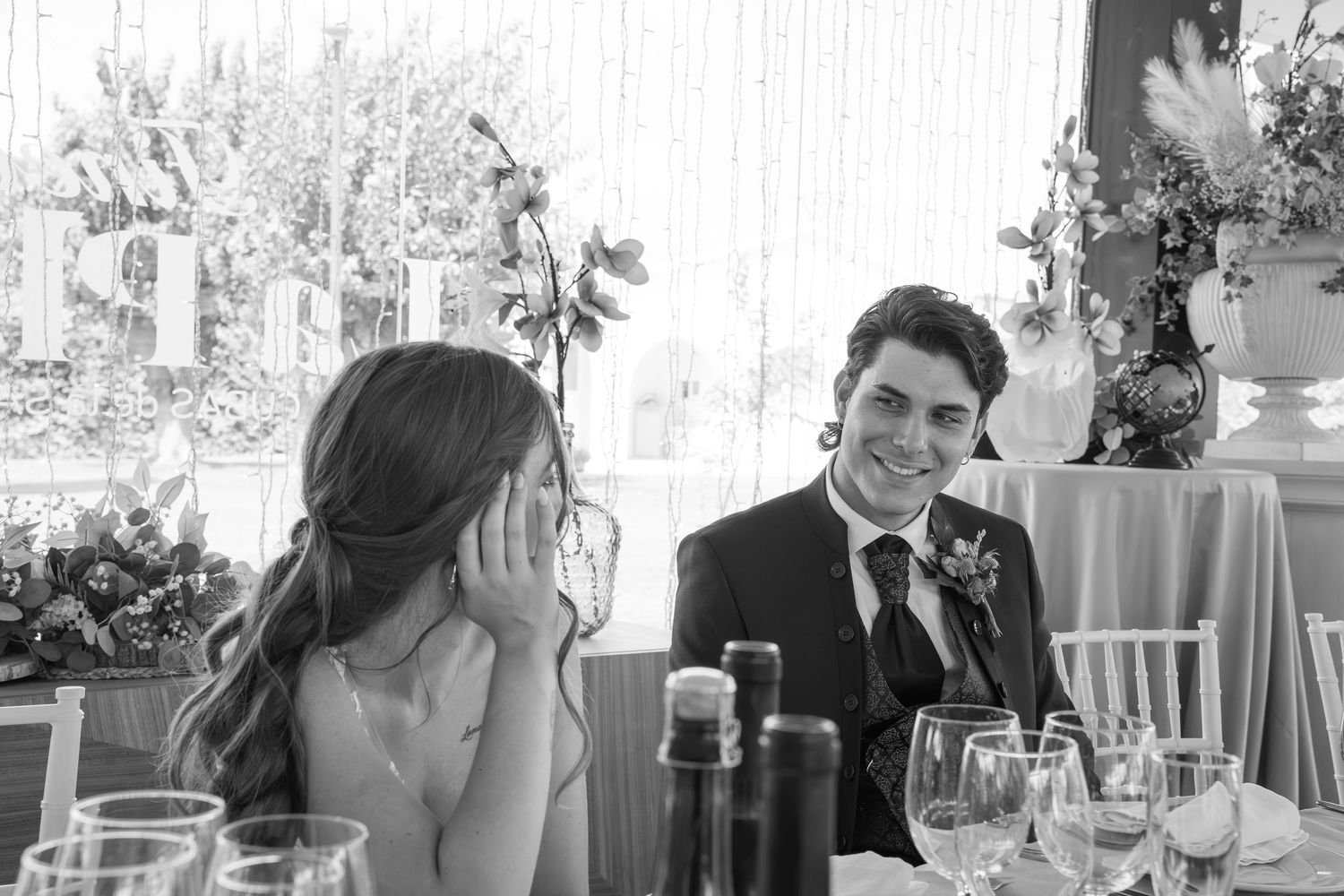 Smiling groom in a dark suit with boutonniere sits at a wedding reception table beside a woman resting her cheek on her hand.
