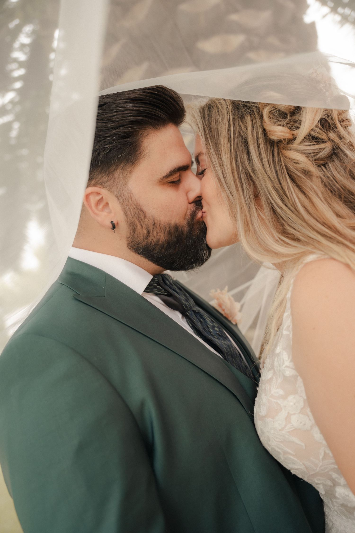 Bride and groom share a kiss beneath a sheer veil, he in a green suit and dark tie, she in a lace wedding gown.