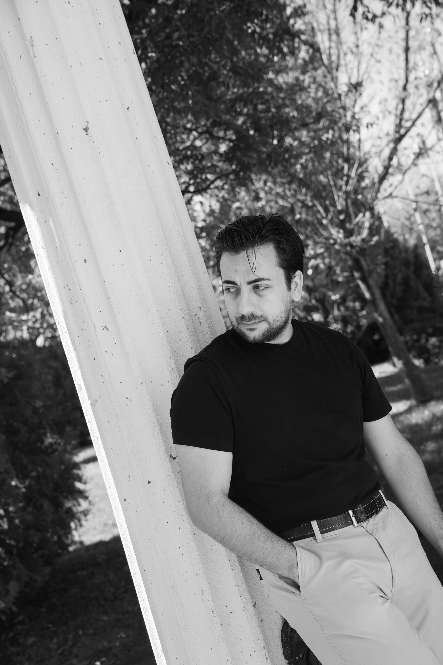 Black-and-white portrait of a bearded man in a black t-shirt and light pants leaning against a white fluted column outdoors near trees.