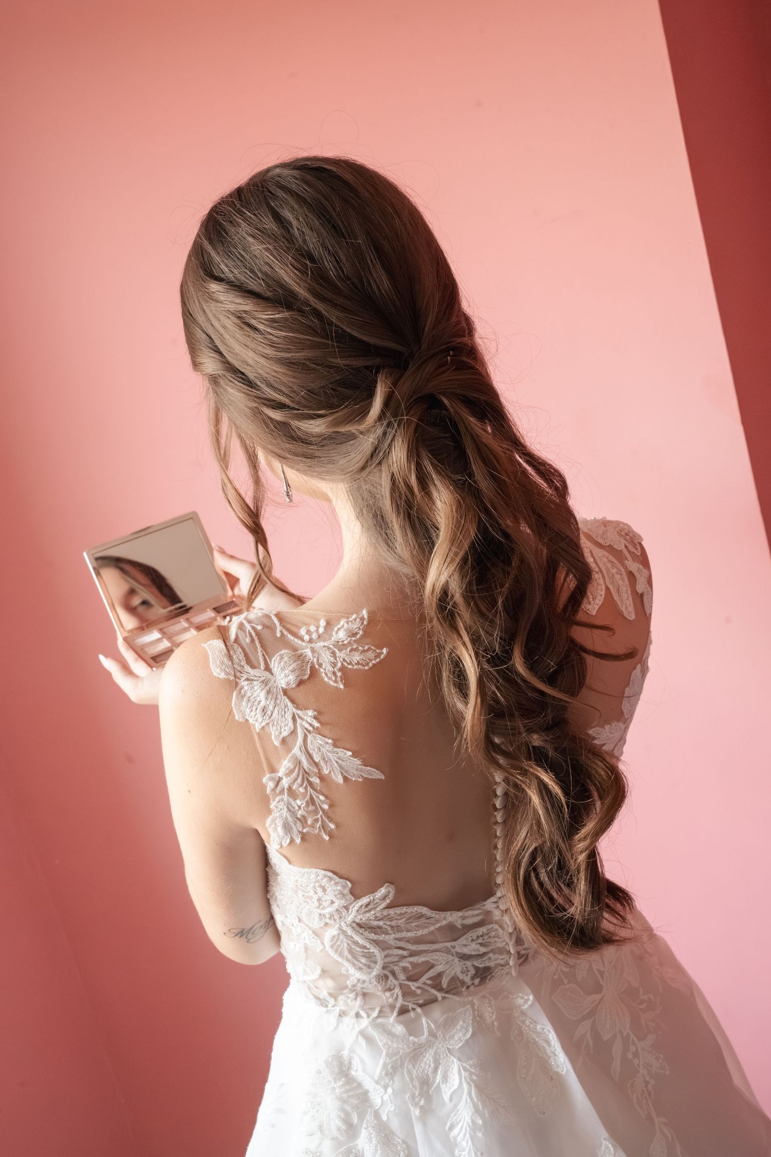 Bride in a white lace wedding gown with floral back details and long wavy half-up hair looks into a compact mirror against a pink wall.