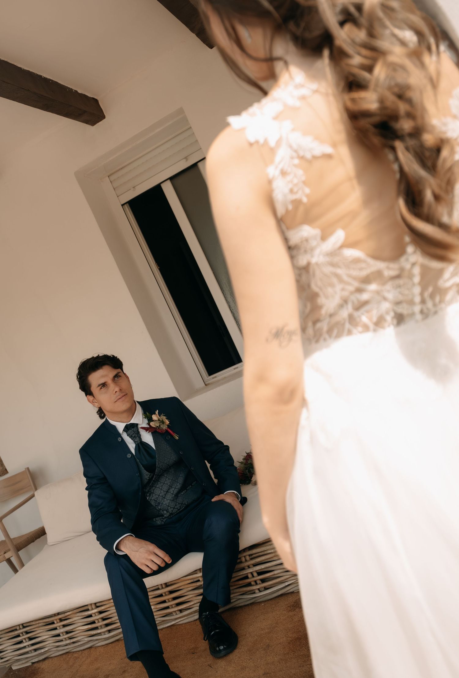 Groom in a navy suit with a boutonniere sits on a wicker sofa, gazing at a bride in a lace-back wedding gown standing in the foreground.