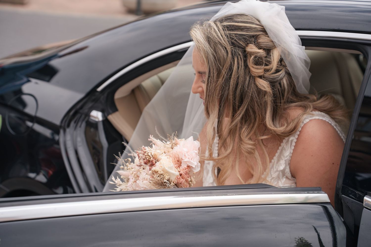 Bride in a white veil and braided updo holding a pink floral bouquet as she steps out of a black car.