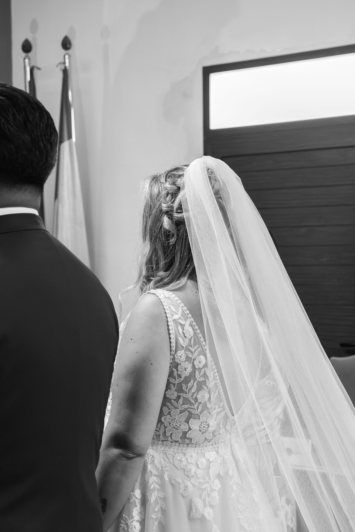 Black and white wedding photo of a bride in a lace gown and long veil standing beside the groom during a ceremony.