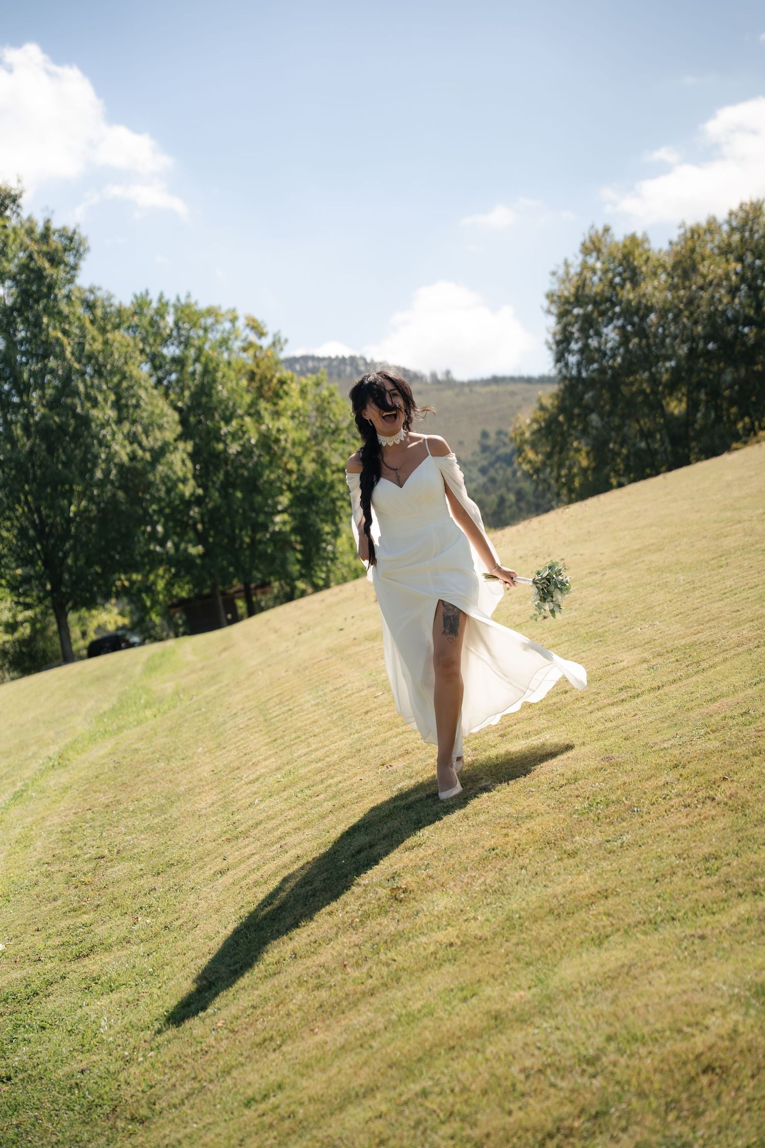 Bride in a white off-the-shoulder wedding dress walks barefoot with a bouquet across a sunny grassy hillside surrounded by green trees.