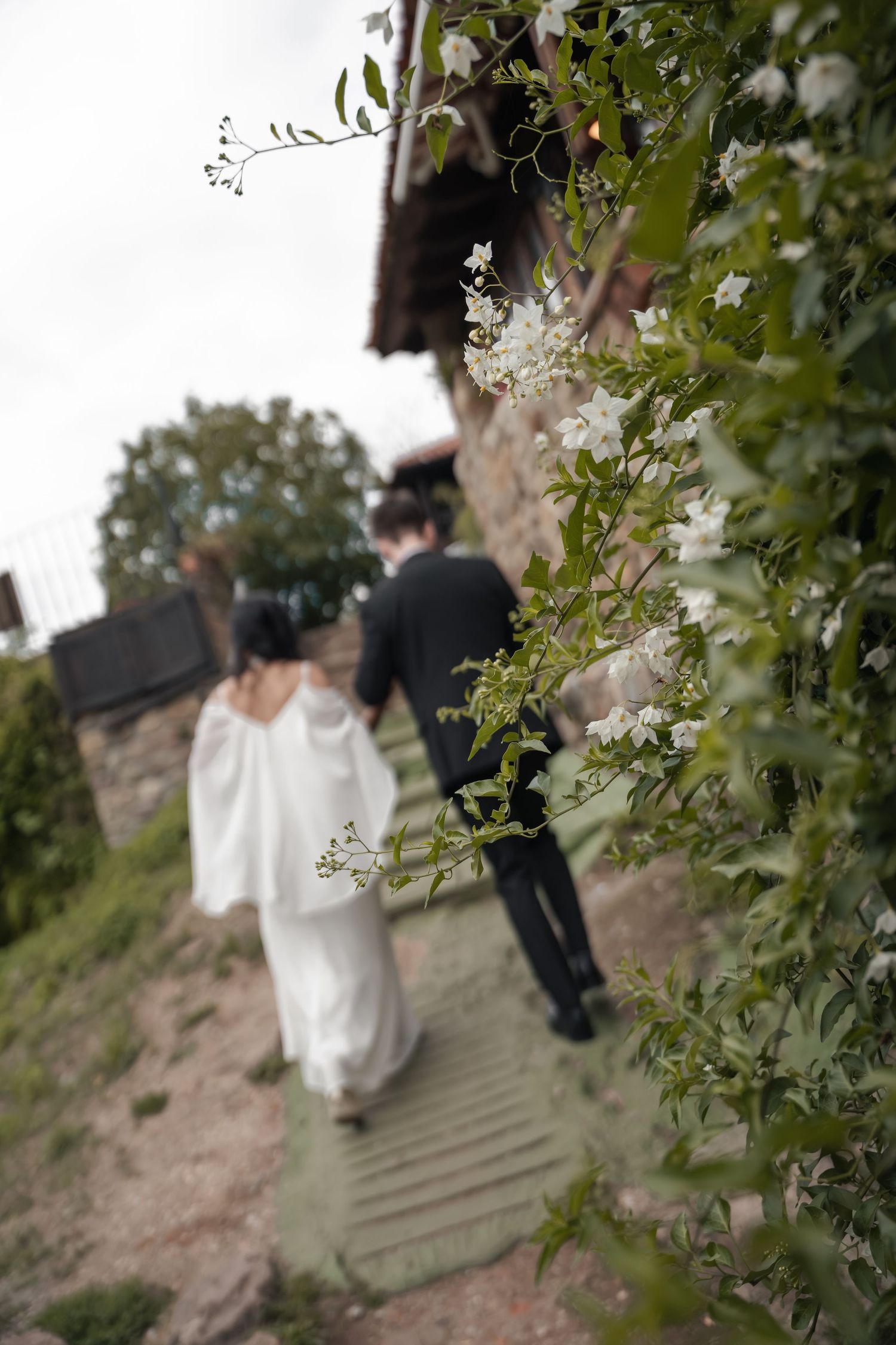 Bride in a white flowing dress and groom in a dark suit walk a stone path past blooming white flowers toward a rustic stone building.