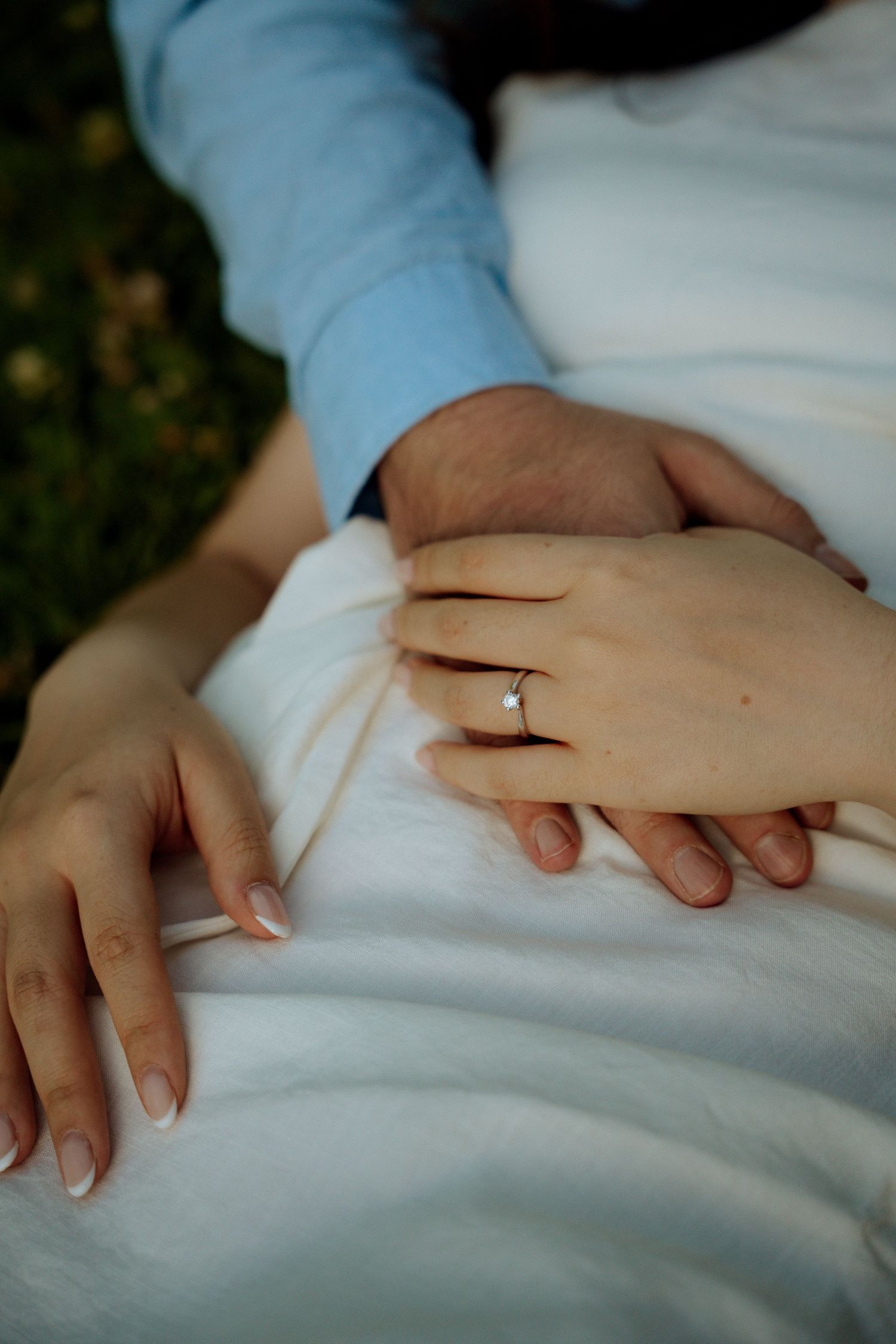 Close-up of couple holding hands with bride wearing a delicate silver engagement ring on white dress.