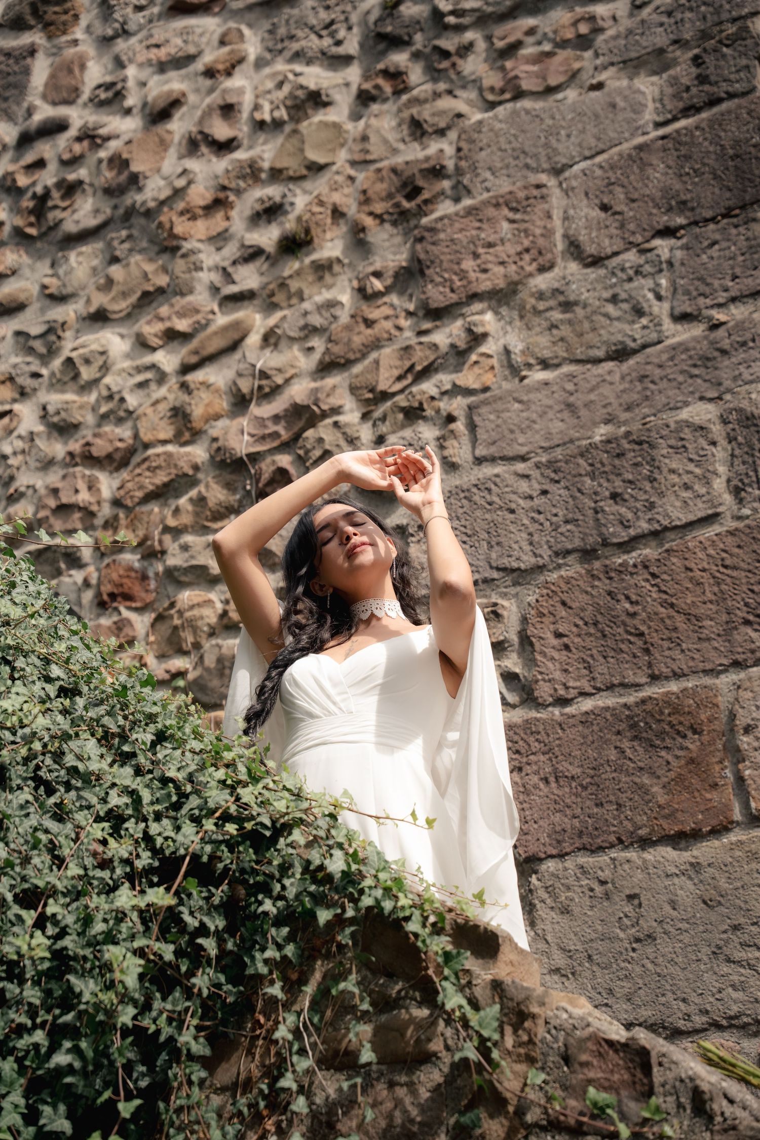Woman in white dress posing gracefully against a rustic stone wall with green ivy, smiling in sunlight.