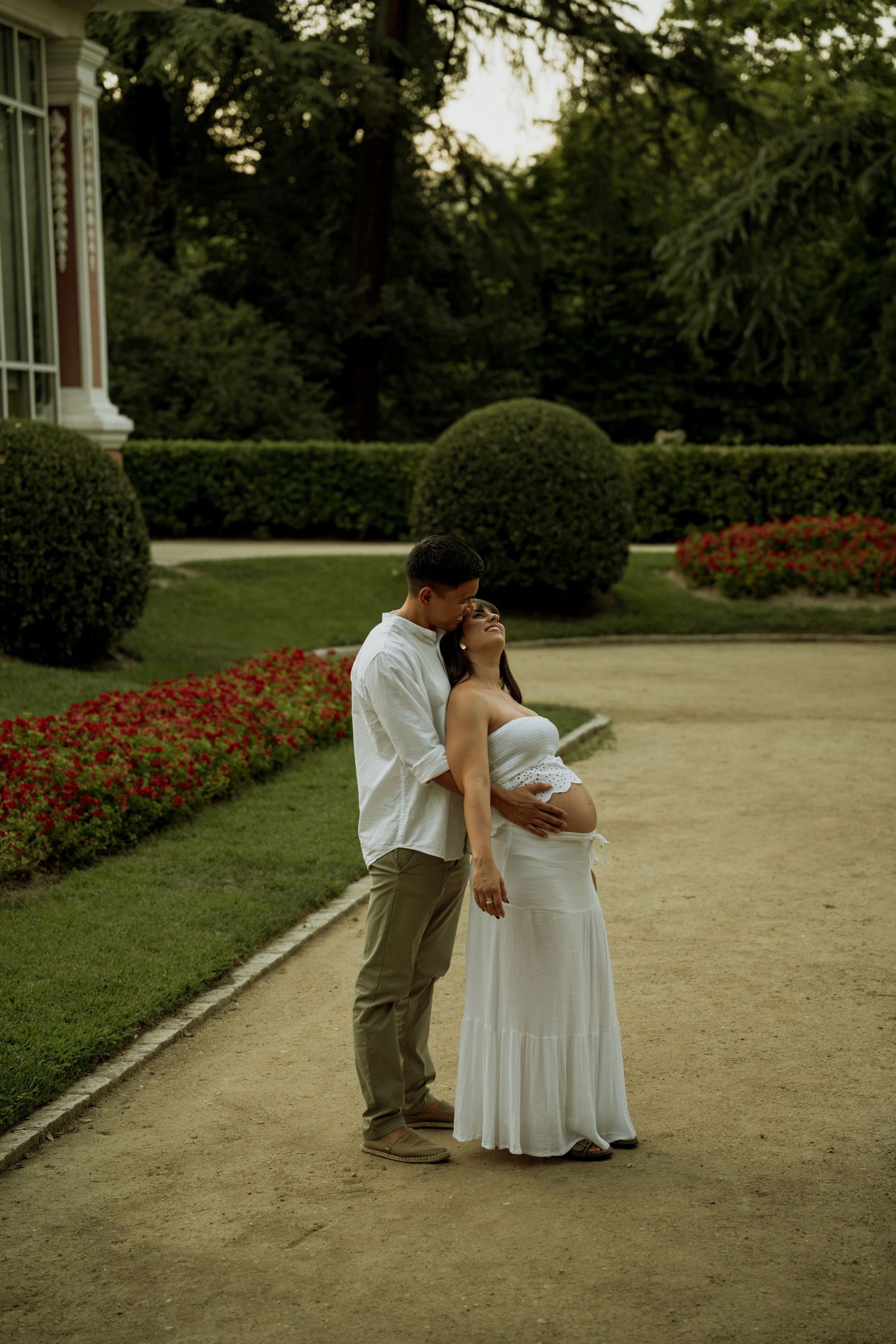 Couple sharing tender moment in garden, pregnant woman in white dress embraced by partner.