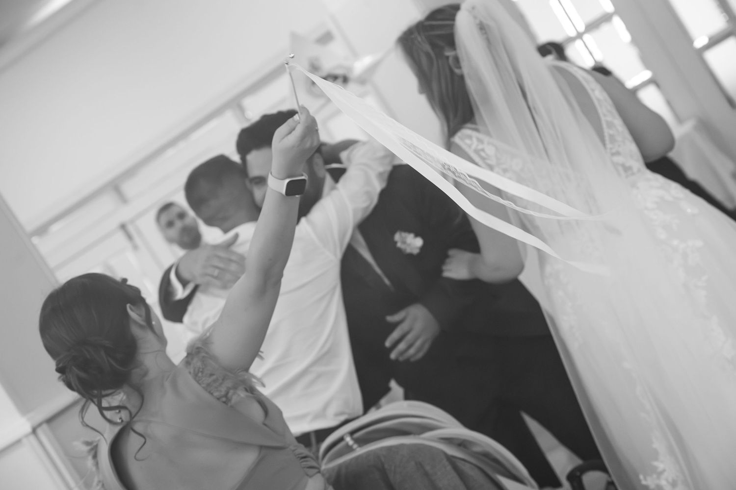 Black and white photo of wedding guests lifting a long bridal veil above a bride in a lace gown during a joyful celebration.