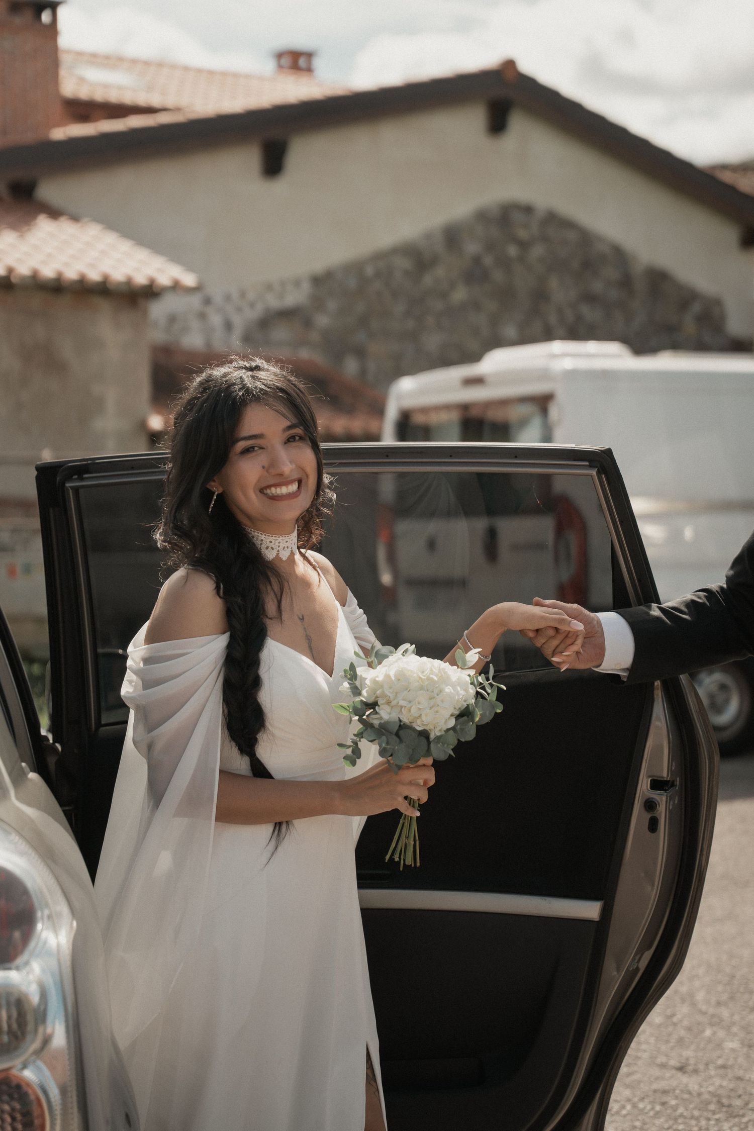 Smiling bride in a white satin gown holding a white floral bouquet as she steps out of a black car and takes a groom's hand.