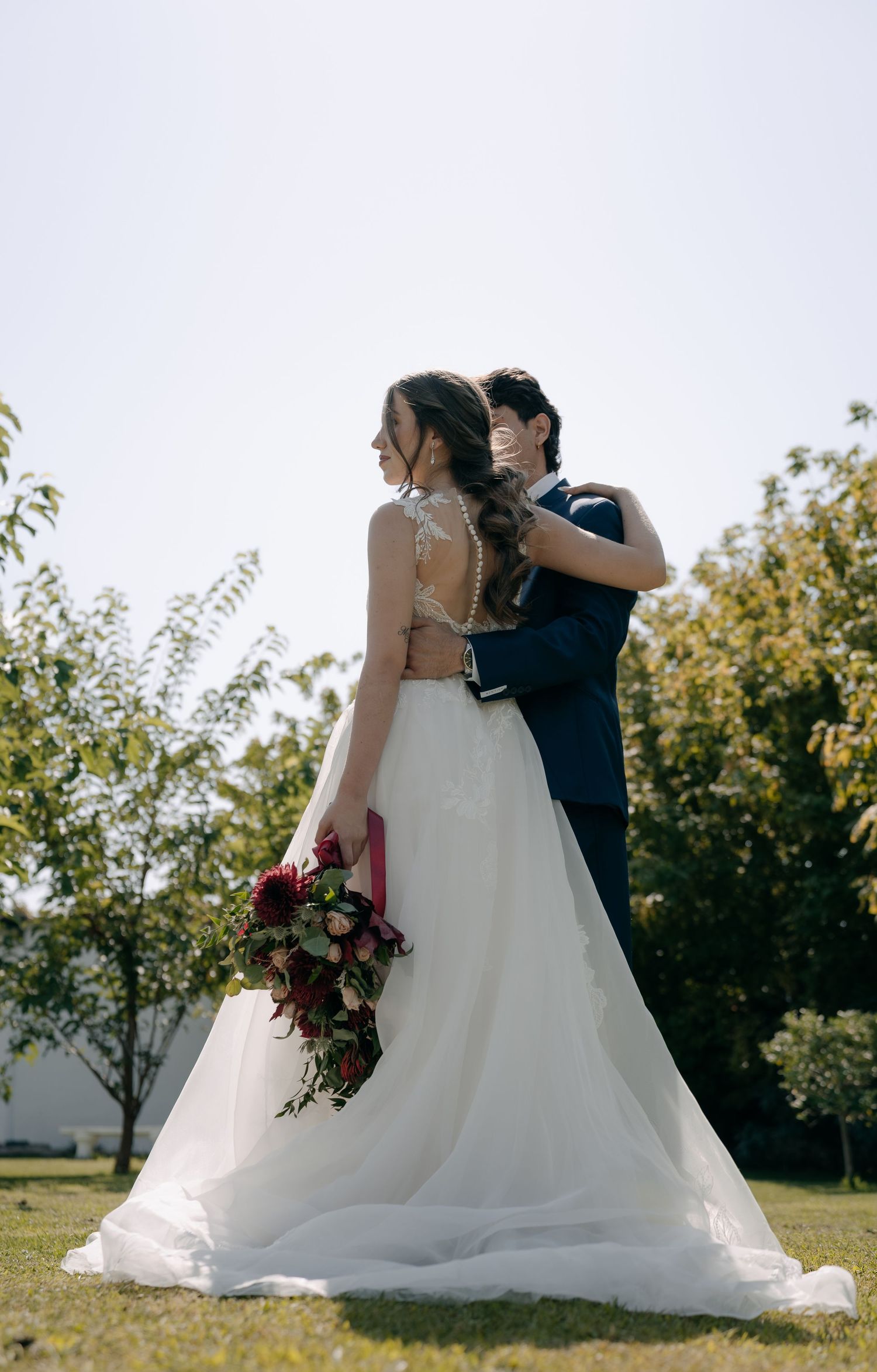 Bride in white gown holding red bouquet embraced by groom in navy suit outdoors on sunny wedding day.