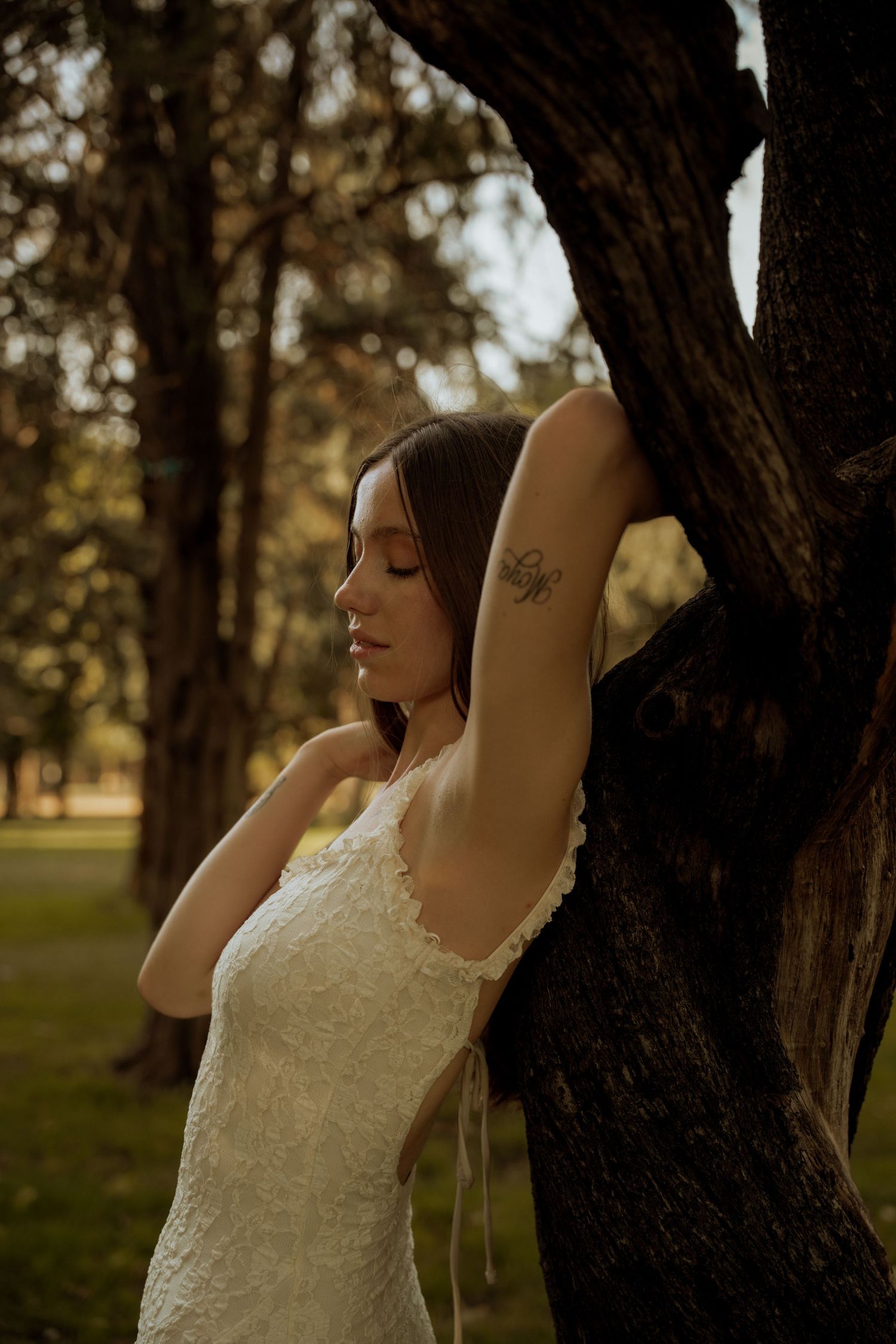 Woman in a white lace dress leans against a tree trunk with her arm raised, showing a small arm tattoo in warm golden park light.