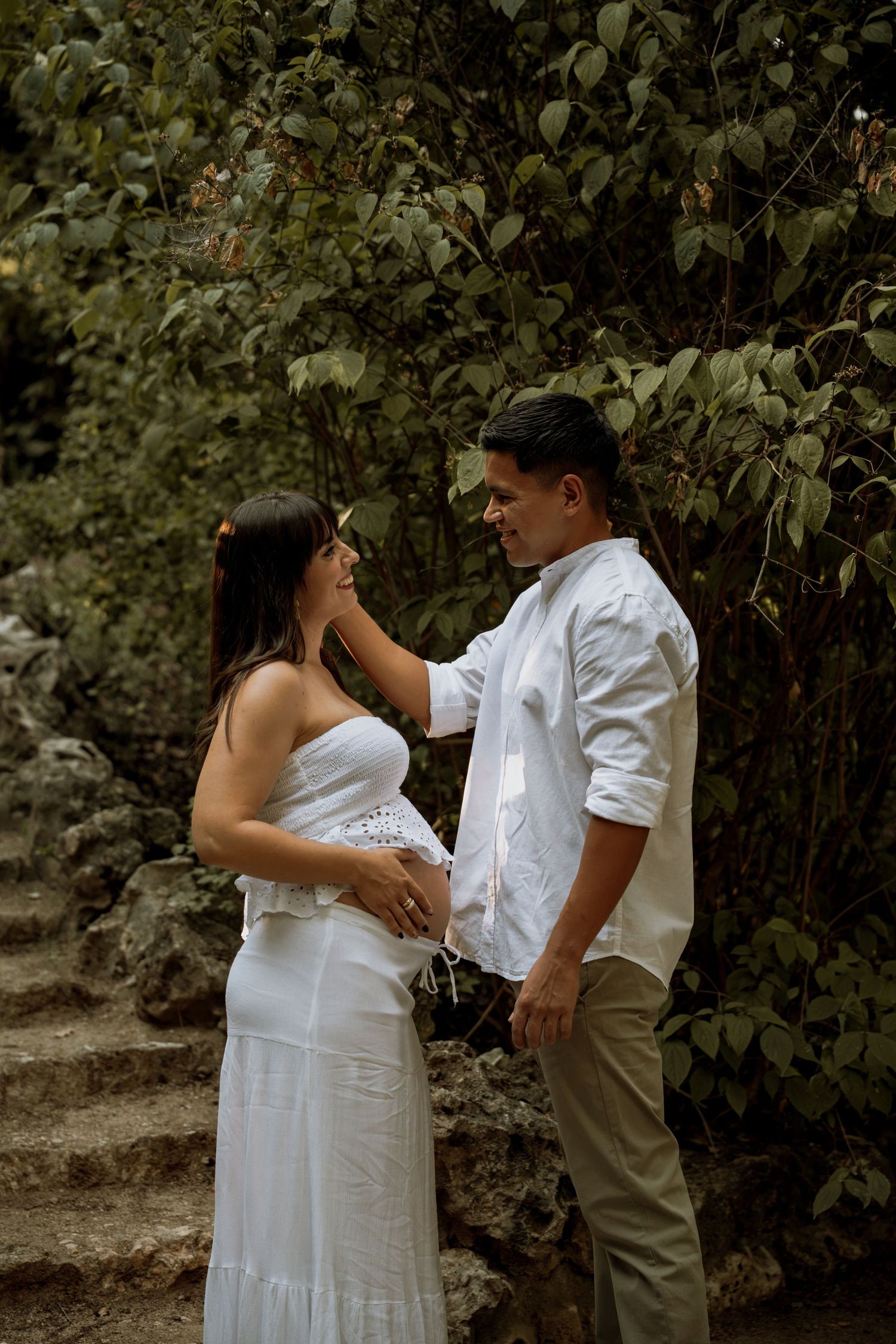 Expecting couple sharing tender moment on stone steps, man gently touching pregnant woman's face outdoors.
