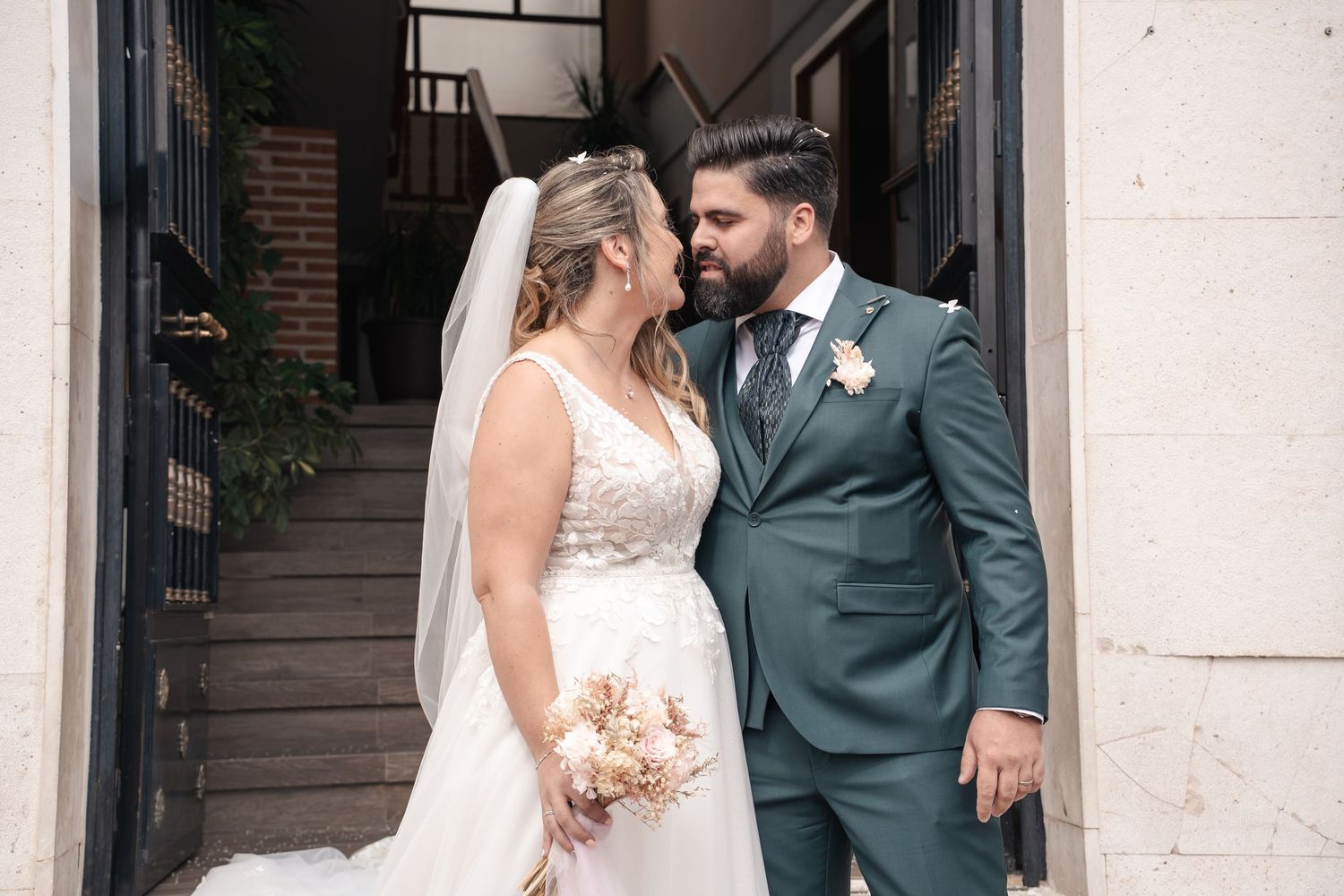 Bride in a lace wedding dress and veil holding a pastel bouquet embraces a bearded groom in a green suit outside a stone building.