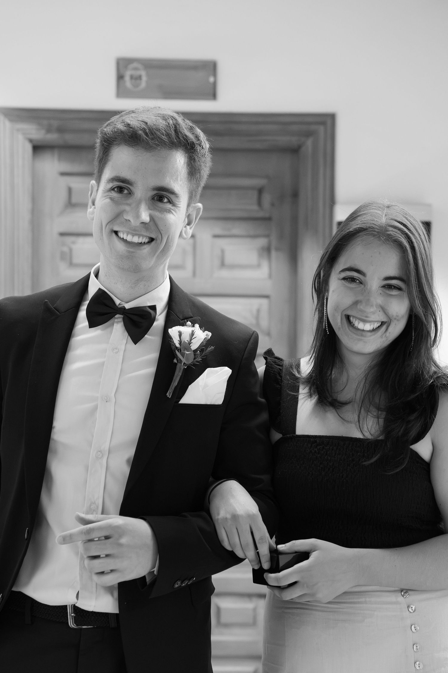 Smiling man in a tuxedo with bow tie and boutonniere stands arm in arm with a smiling woman in a dark dress at a formal event.