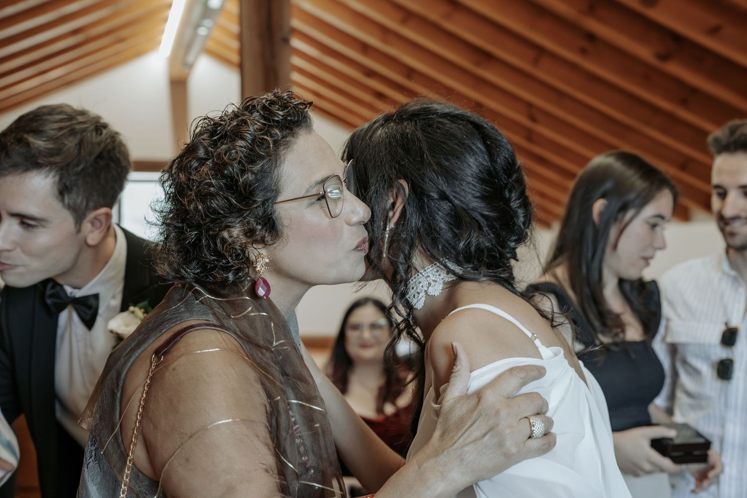 A curly-haired woman in glasses kisses a woman in a white dress on the cheek as wedding guests smile in the background.