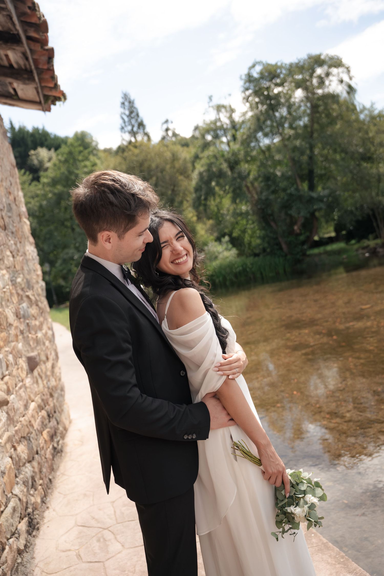 A smiling bride and groom embrace on a stone path beside a calm pond, with lush green trees and a bridal bouquet in her hand.