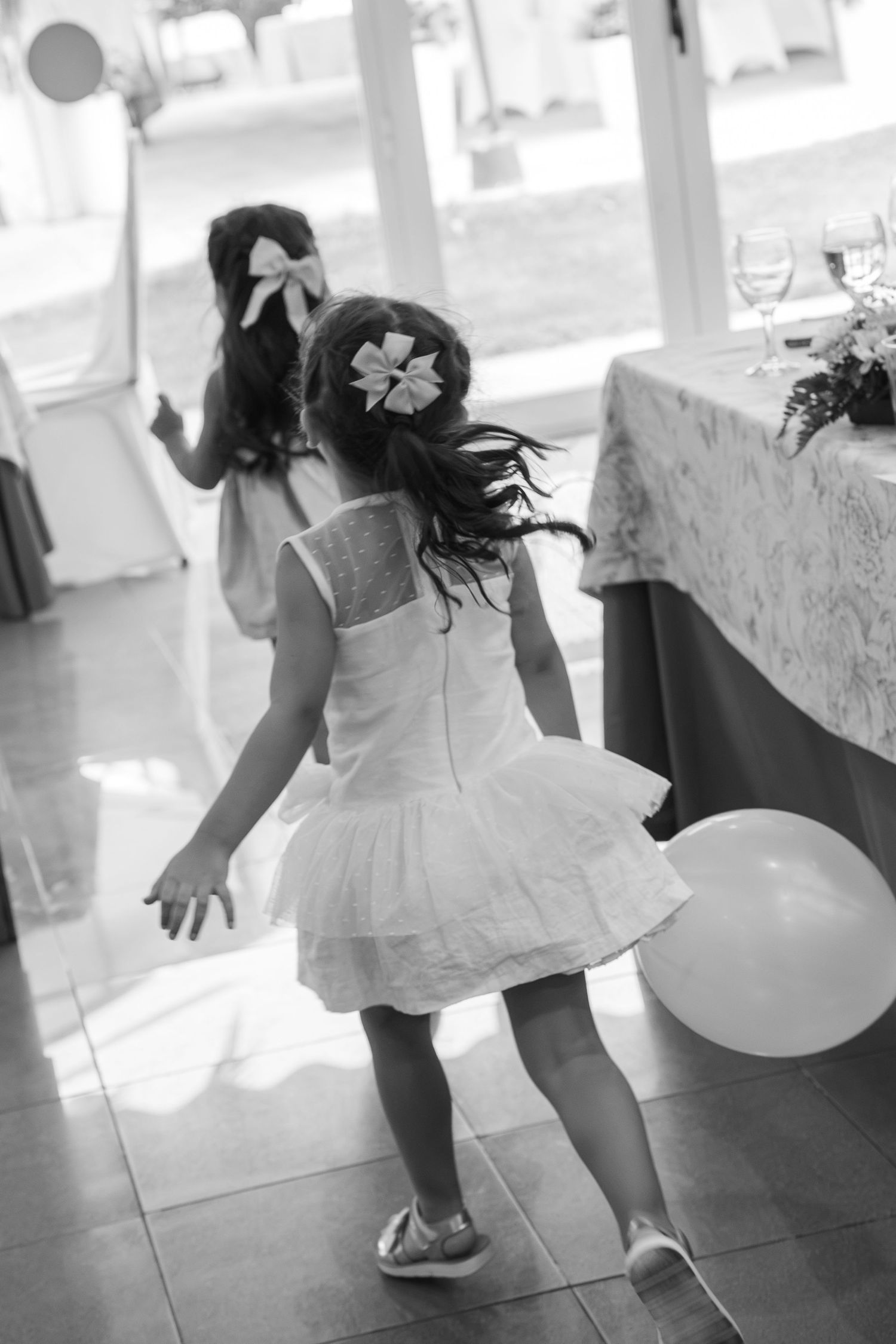 Two little girls in white dresses and hair bows dance and play near a white balloon at a wedding reception in black and white.