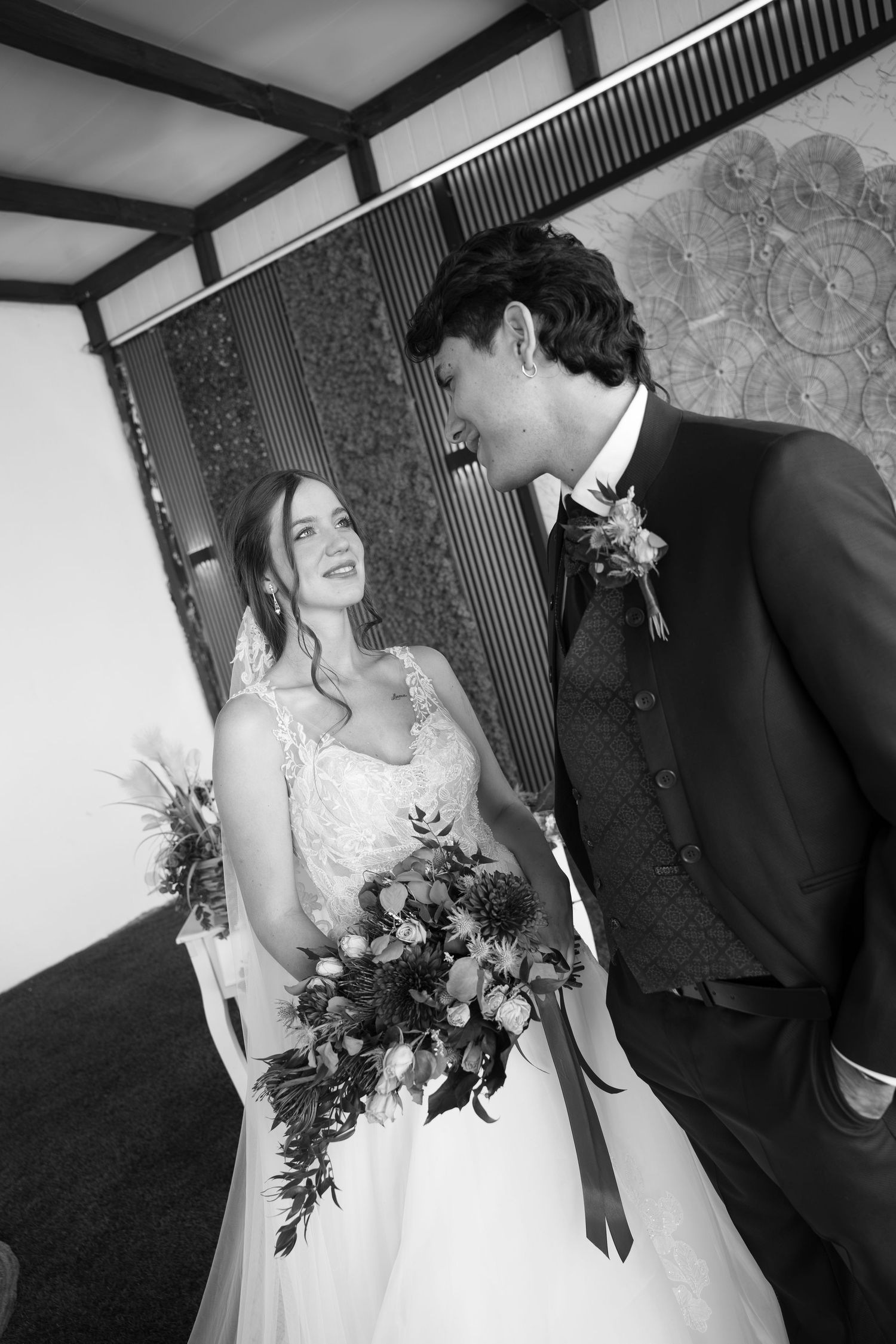 Black and white photo of a smiling bride in a lace gown holding a bouquet as the groom in a suit and boutonniere gazes at her.