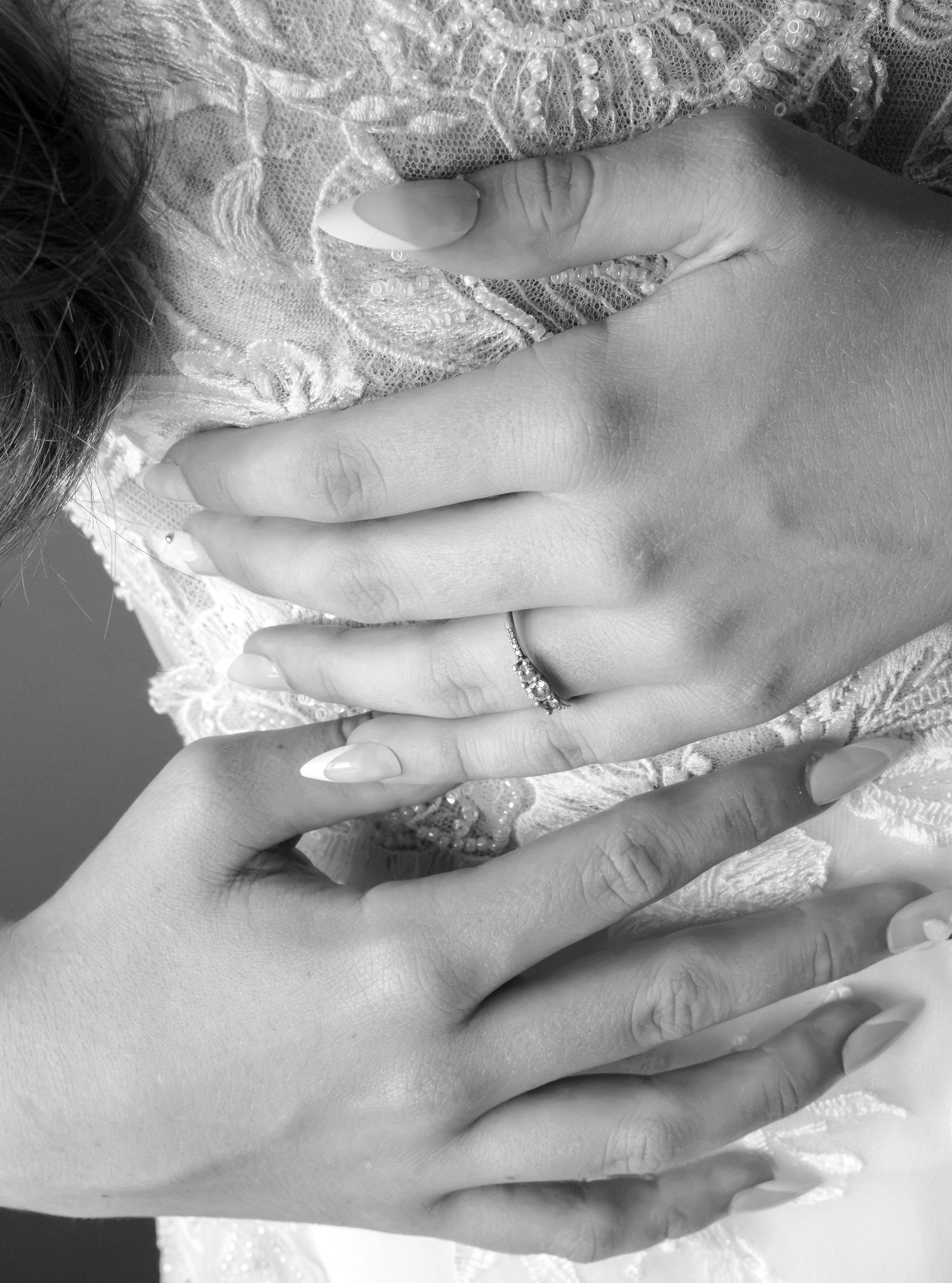 Black and white close-up of a bride's hands clasped over a lace wedding dress, showing a sparkling diamond engagement ring.