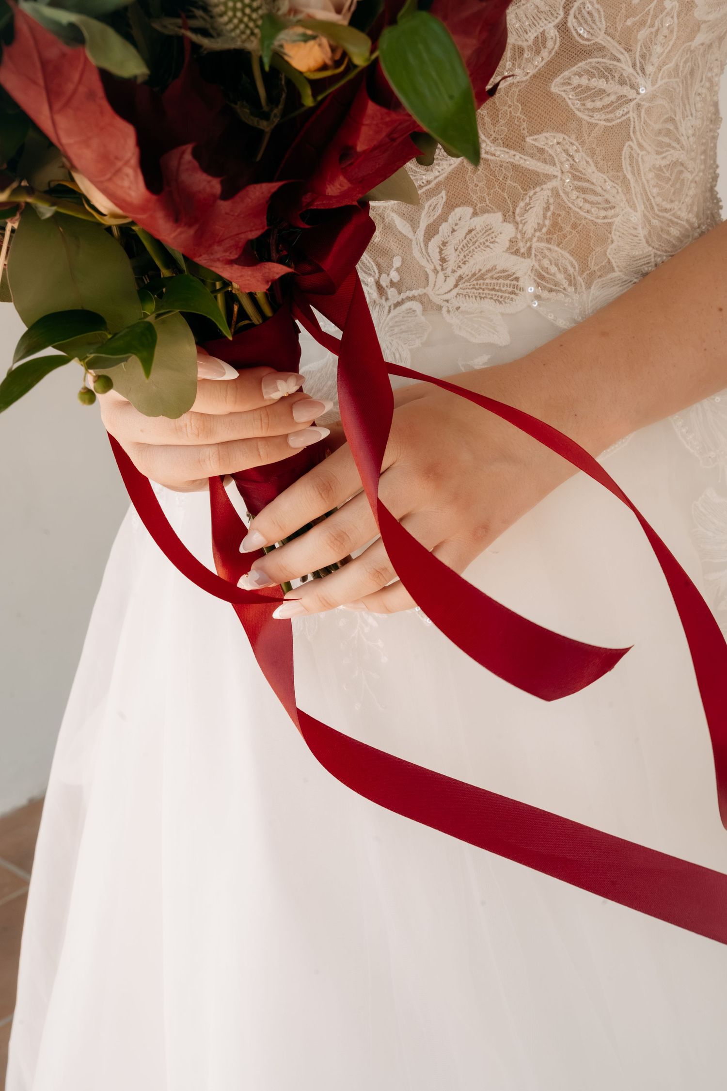 Bride in a white lace and tulle wedding gown holding a bouquet of burgundy flowers and green leaves tied with long red satin ribbons.