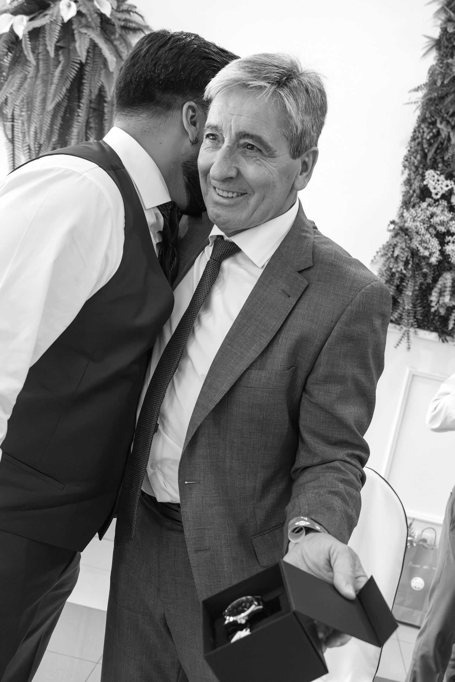 Smiling man in a gray blazer holds an open gift box with a wristwatch while another man in a vest whispers in his ear.