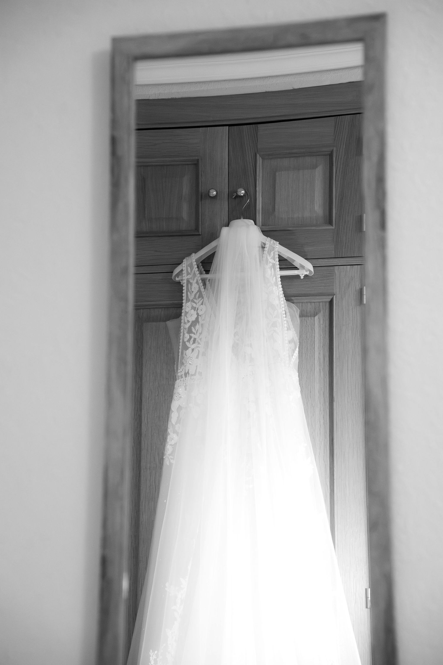 Black and white photo of a lace wedding dress with a long veil hanging on a hanger against a wooden door, seen in a mirror.