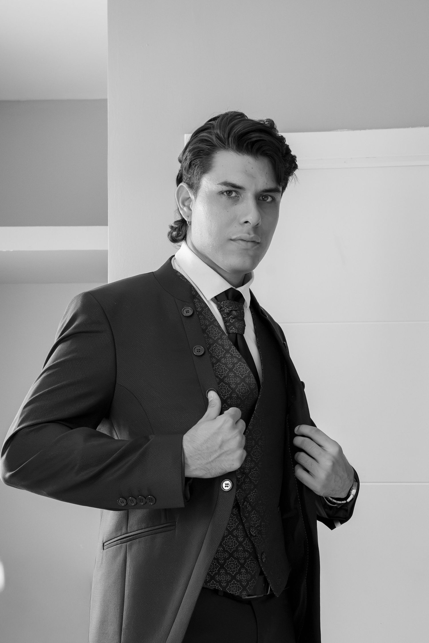 Black and white portrait of a young man in a tailored suit and patterned vest, adjusting his jacket lapel.