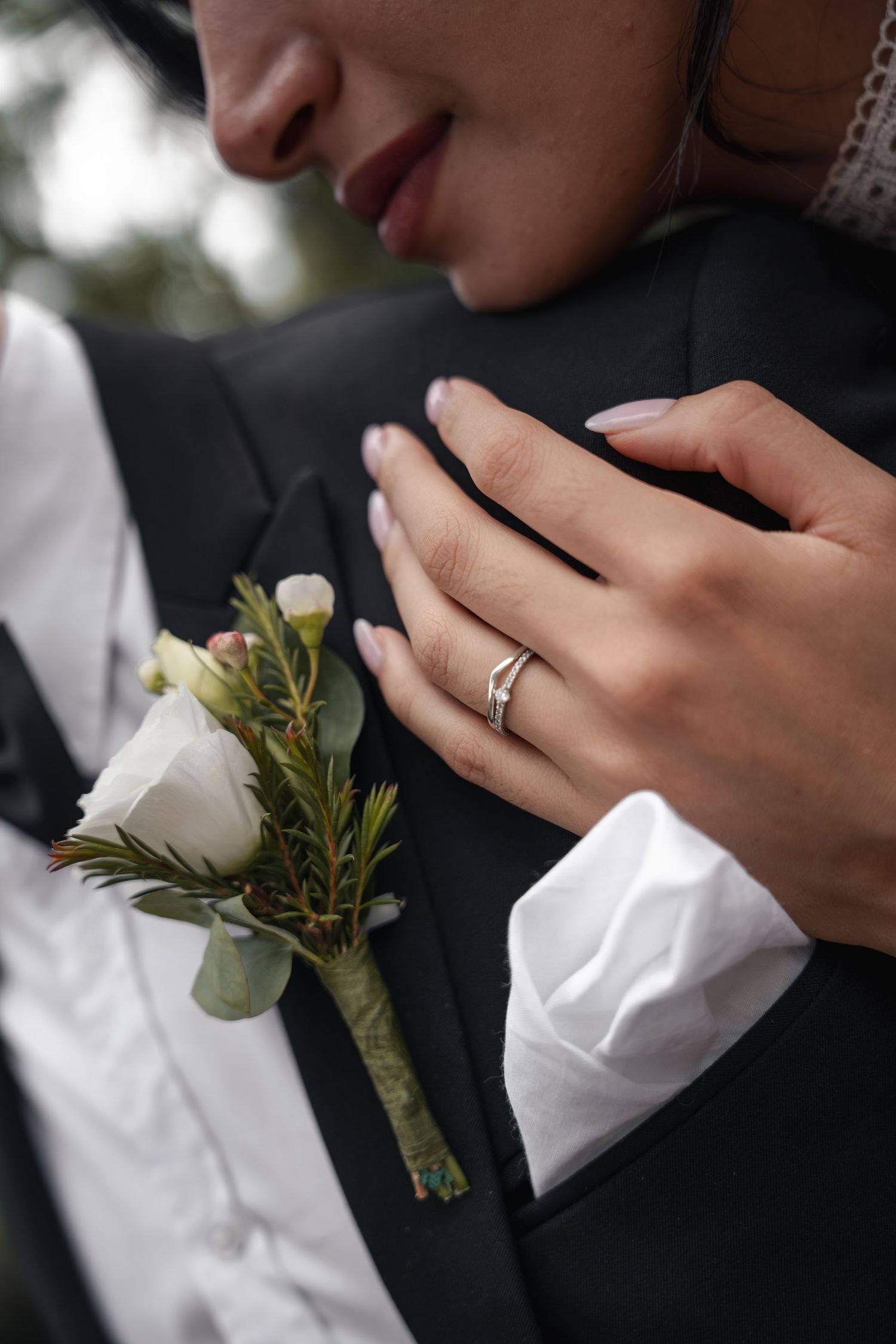 Close-up of bride wearing diamond ring resting hand on groom's black suit lapel with white rose boutonniere.