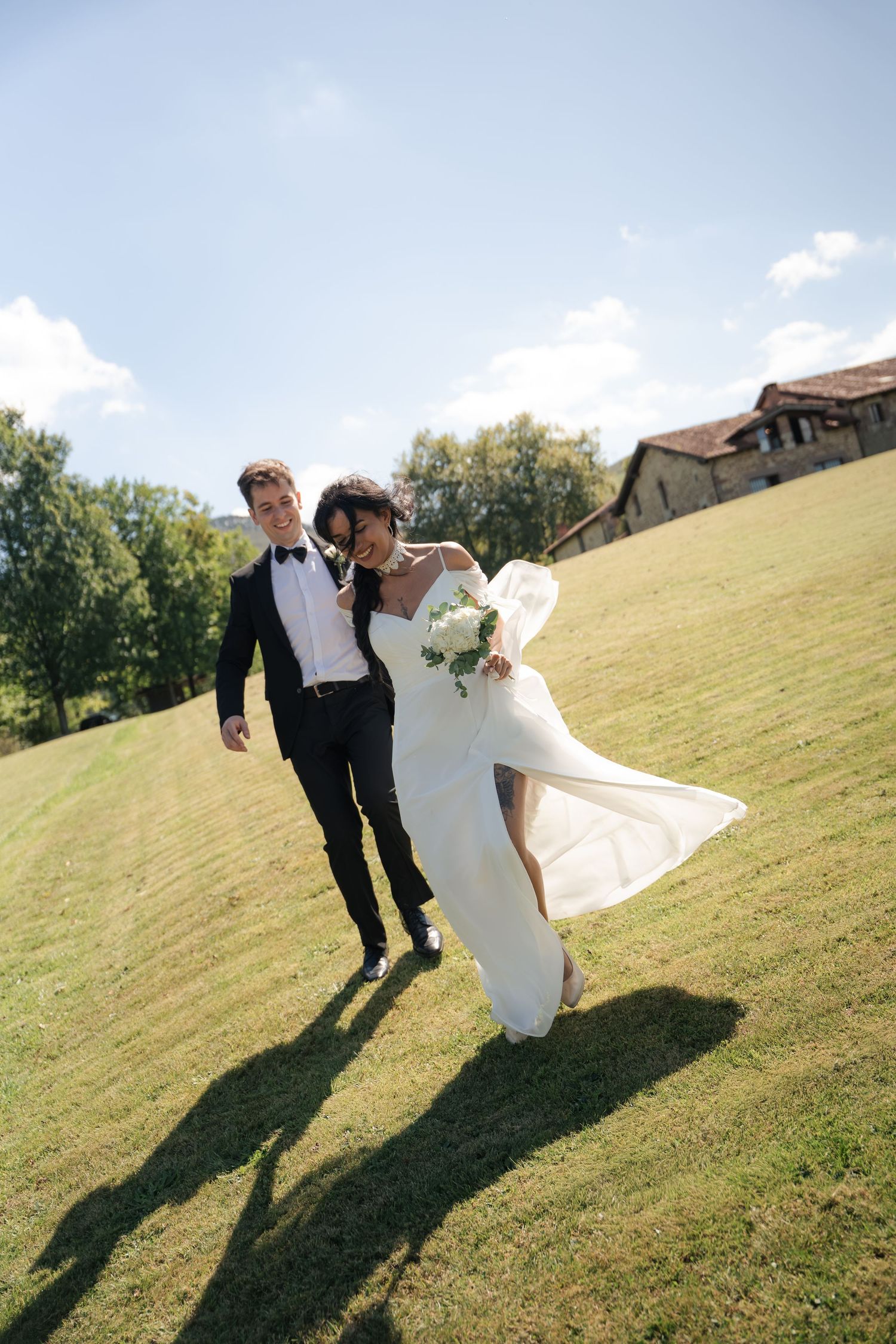 Joyful newlyweds walking on green grass, bride in white gown holding bouquet, groom in black tuxedo, sunny outdoor wedding.